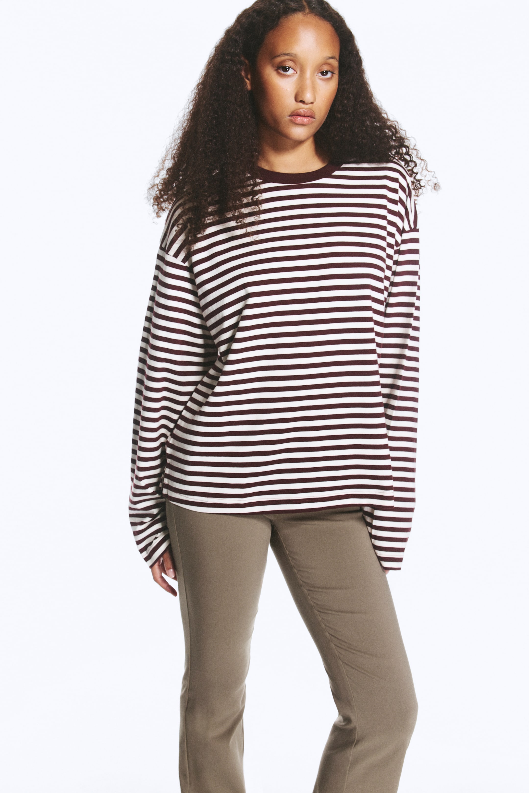 Agrandir l'image: Le mannequin porte un T-shirt à manches longues coupe décontractée, à rayures horizontales bordeaux et blanches, avec des épaules tombantes et un col rond bordeaux uni. Le T-shirt est porté avec un pantalon vert olive.