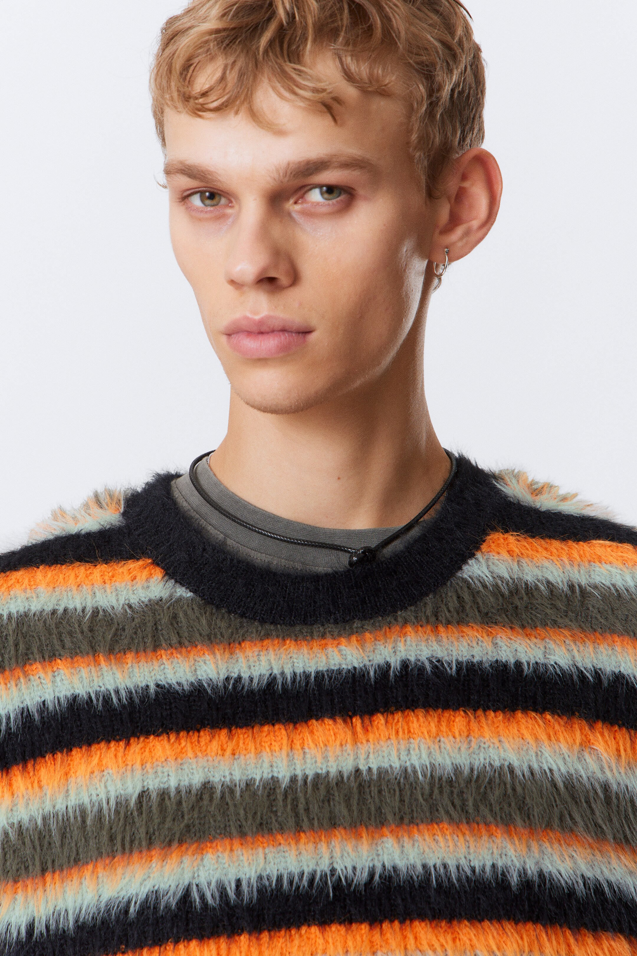Se større billede: En strikket pullover med rund hals og fyldig struktur vises forfra, med horisontale striber i sort, skrigende orange, lys blå og olivengrøn. Den er lagdelt over en grå t-shirt, med en tynd sort snor halskæde synlig under sweaterens halsudskæring.