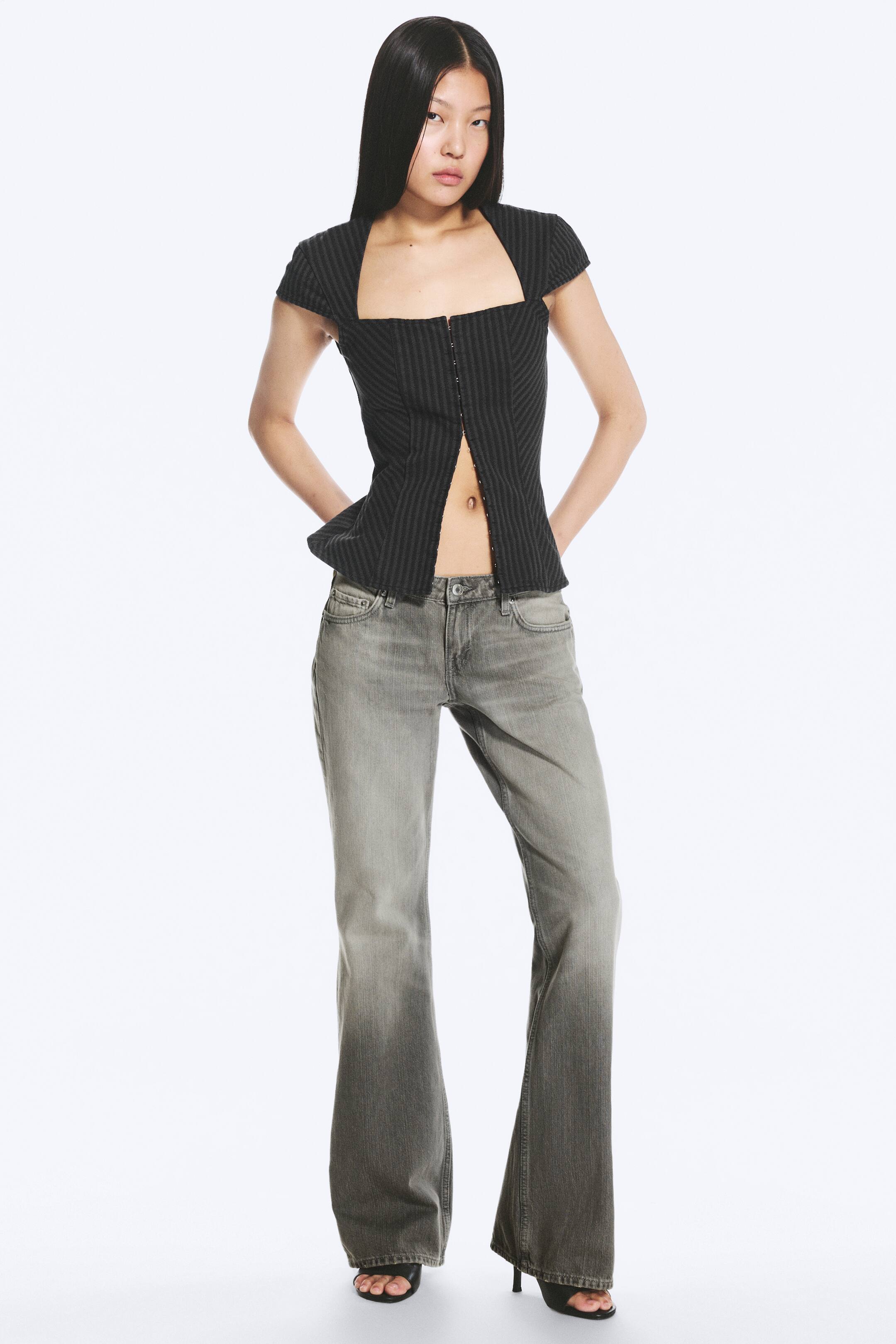 Grotere afbeelding bekijken: Grijze low-rise jeans met een slim fit en bootcut pijp worden gedragen door het model. De jeans hebben een verwassen denimlook met strepen, een klassiek 5-pocketdesign en een ritssluiting, en vallen lang waardoor de schoenen van het model gedeeltelijk bedekt worden.