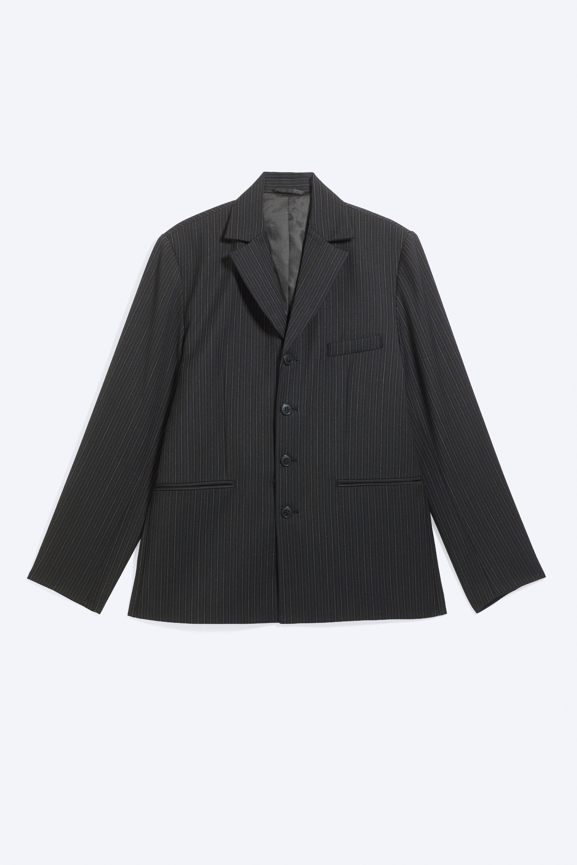 La modelo luce una americana de mezcla de lana, cruzada sencilla y con finas rayas oscuras, mientras posa de pie con los brazos cruzados. La americana presenta cuello alzado y se combina con un pantalÃ³n de pinstripes a juego, una camisa oscura y una capa de punto de tono claro visible en las mangas remangadas.