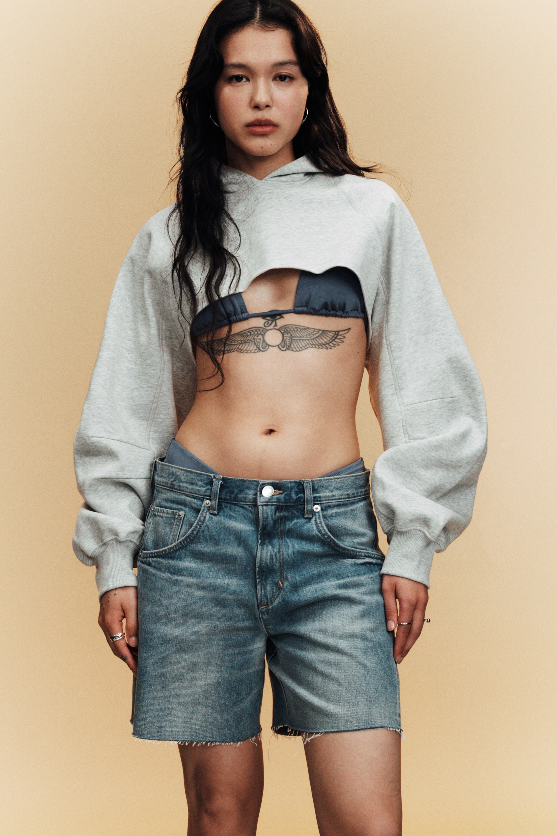 Ver imagen más grande: Una sudadera con capucha gris recortada presenta un dobladillo cortado al ras y un gran recorte en el pecho. Está diseñada con mangas largas voluminosas que terminan en puños acanalados, y una capucha visible en la parte trasera.