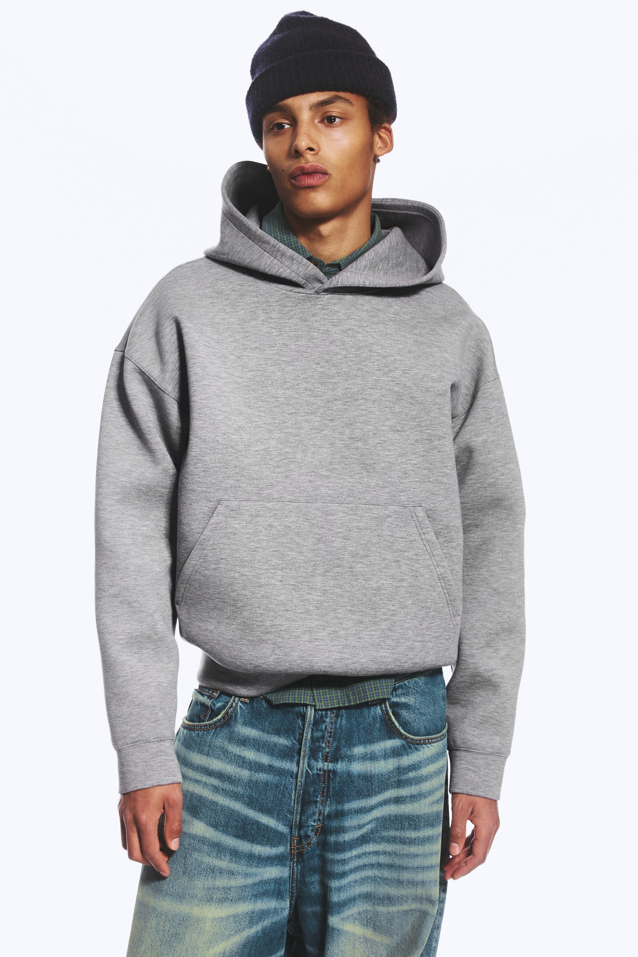 Grotere afbeelding bekijken: Een lichtgrijze scuba hoodie met losse pasvorm, verlaagde schouders en een kangoeroezak aan de voorkant wordt gedragen door het model. De hoodie is gelaagd over een groen hemd met patroon en gestyled met een blauwe jeans en een donker gebreide beanie.