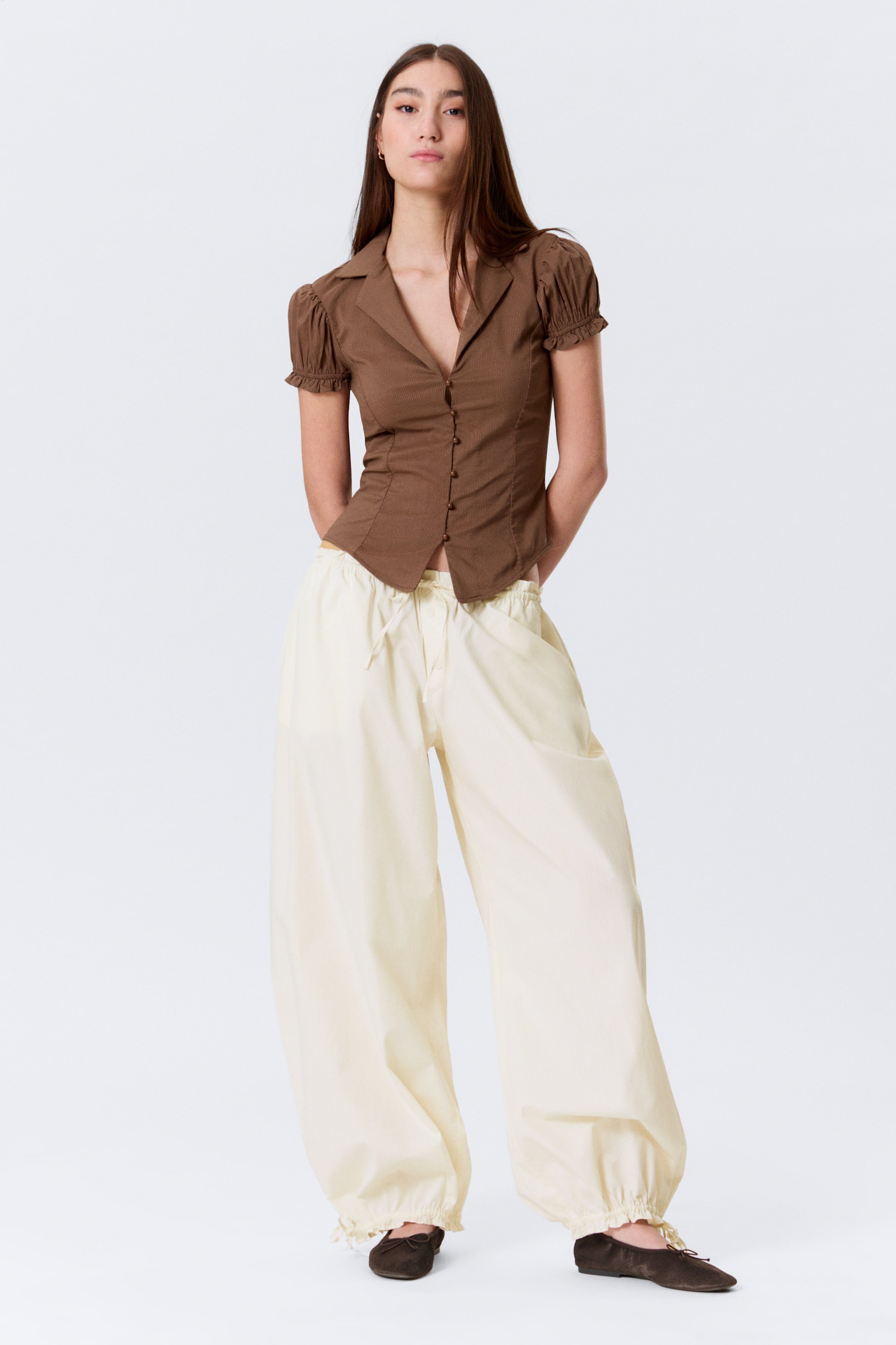 Größeres Bild ansehen: Ein braunes Hemd mit Kragen präsentiert ein figurbetontes Oberteil mit gerafften Details, gepufften kurzen Ärmeln mit gerüschten Manschetten und einem Knopfverschluss vorne. Cremefarbene Hosen mit weitem Bein zeigen einen Kordelzug in der Taille, voluminöse Beine und geraffte Knöchelbündchen.