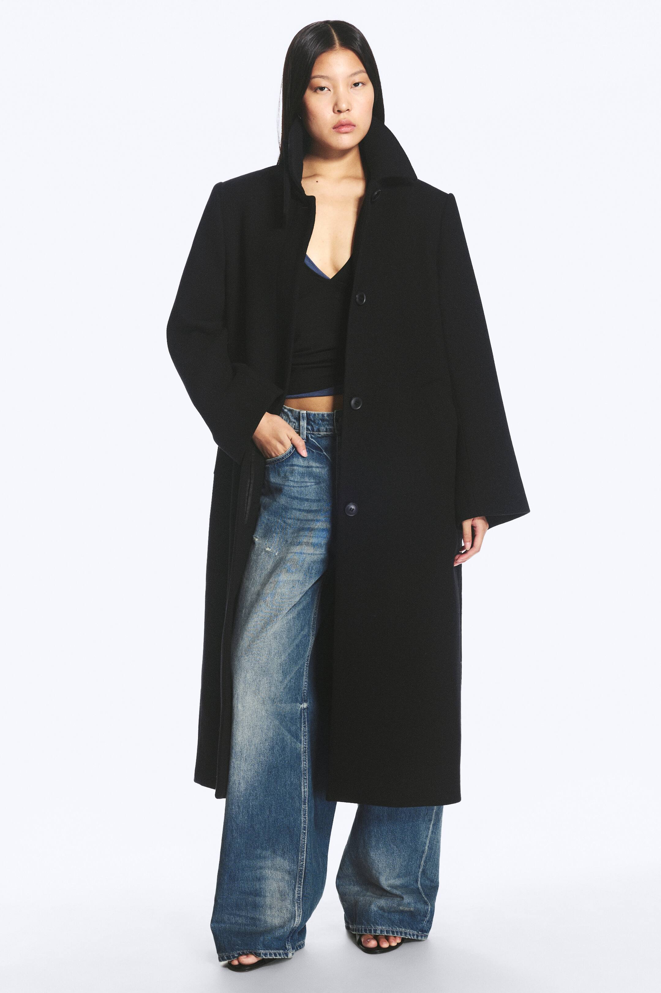 View larger image: The model is wearing a black, long, oversized single-breasted coat. The coat features a turn-down collar and visible front buttons, with the model’s left hand resting in a side pocket.