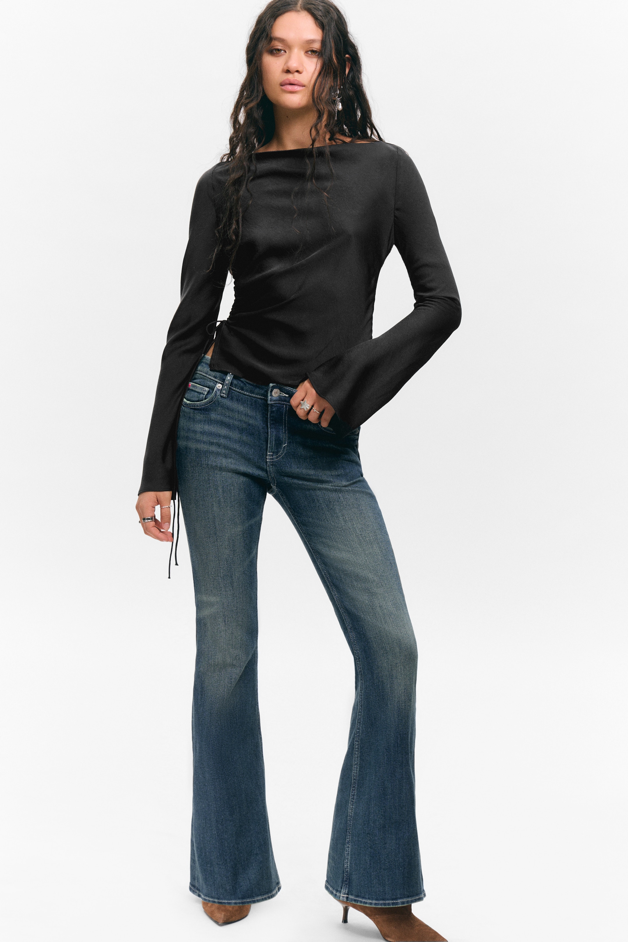 View larger image: A black lustrous long-sleeved top is showcasing a boat neckline and bell sleeves, with functional ties cinching the fabric on the left side. It is styled with mid-wash blue flared jeans, which are closely fitting through the thigh and flaring from the knee, all facing slightly to the right.