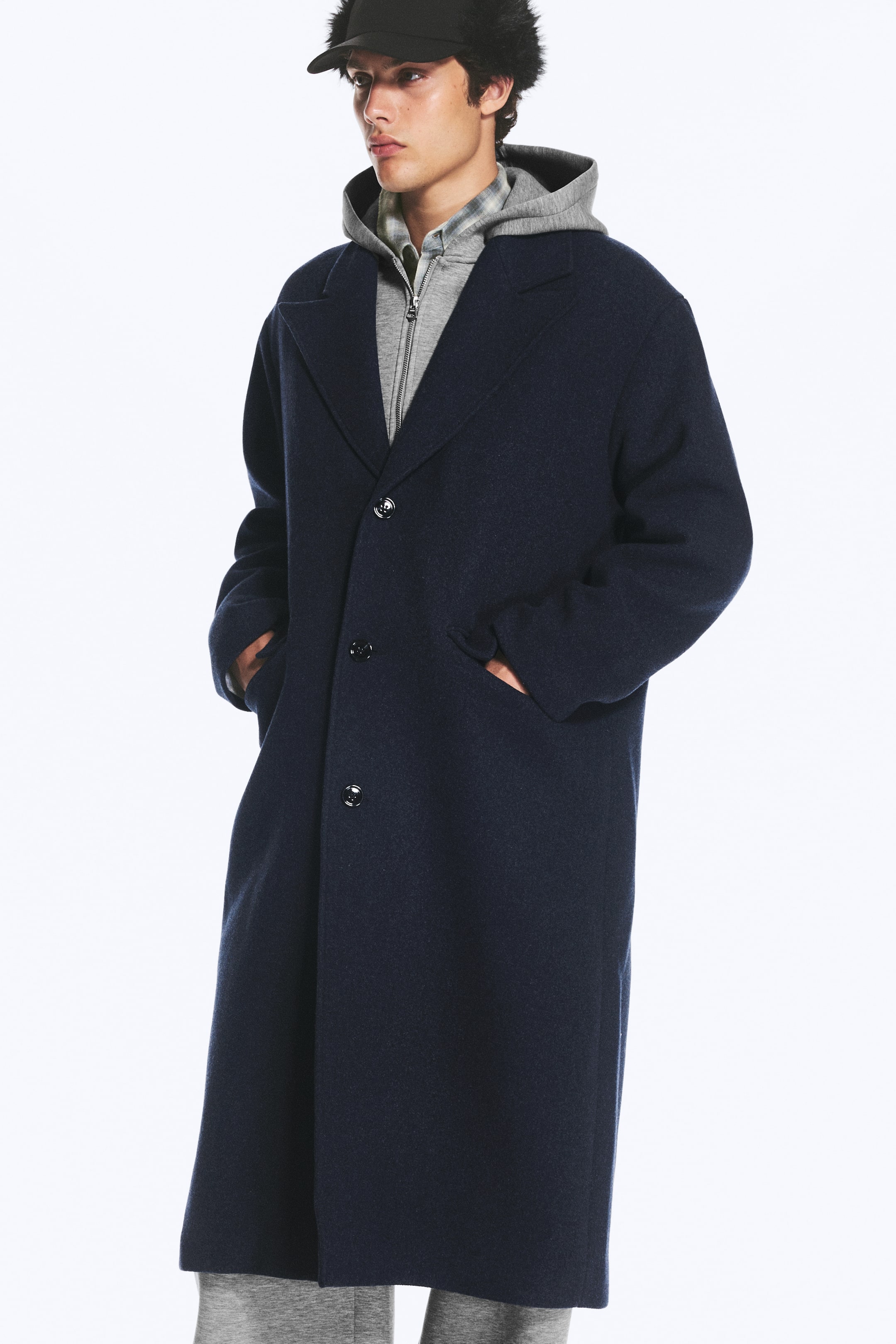 Visualizza immagine più grande: Un lungo cappotto monopetto blu navy con silhouette oversize e revers classico è indossato dalla modella. Il cappotto presenta tre bottoni frontali e tasche laterali, in cui la modella appoggia le mani mentre indossa il capo sopra una felpa con cappuccio grigia con zip.