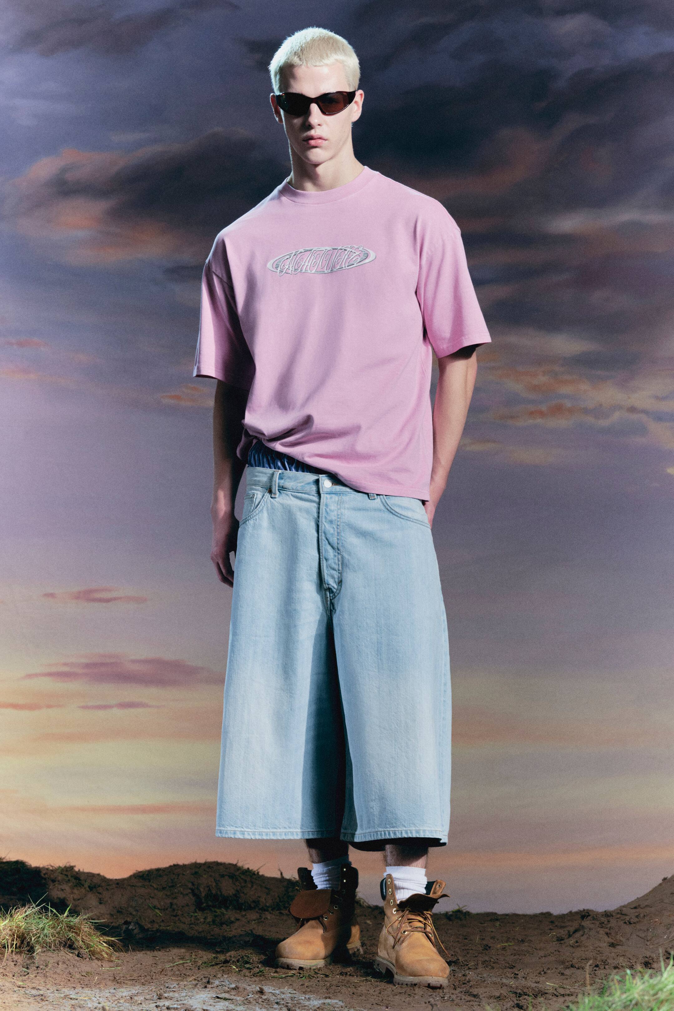 Zobrazit větší obrázek: Růžové oversized tričko s oválným stříbrným iridescentním potiskem je nošeno v kombinaci se světle modrými širokými džínovými kalhotami, které odhalují modrý pruhovaný pas boxerových trenýrek. Světle hnědé pracovní boty na šněrování s bílými ponožkami jsou viditelné a tmavé oválné sluneční brýle spočívají na obličeji.