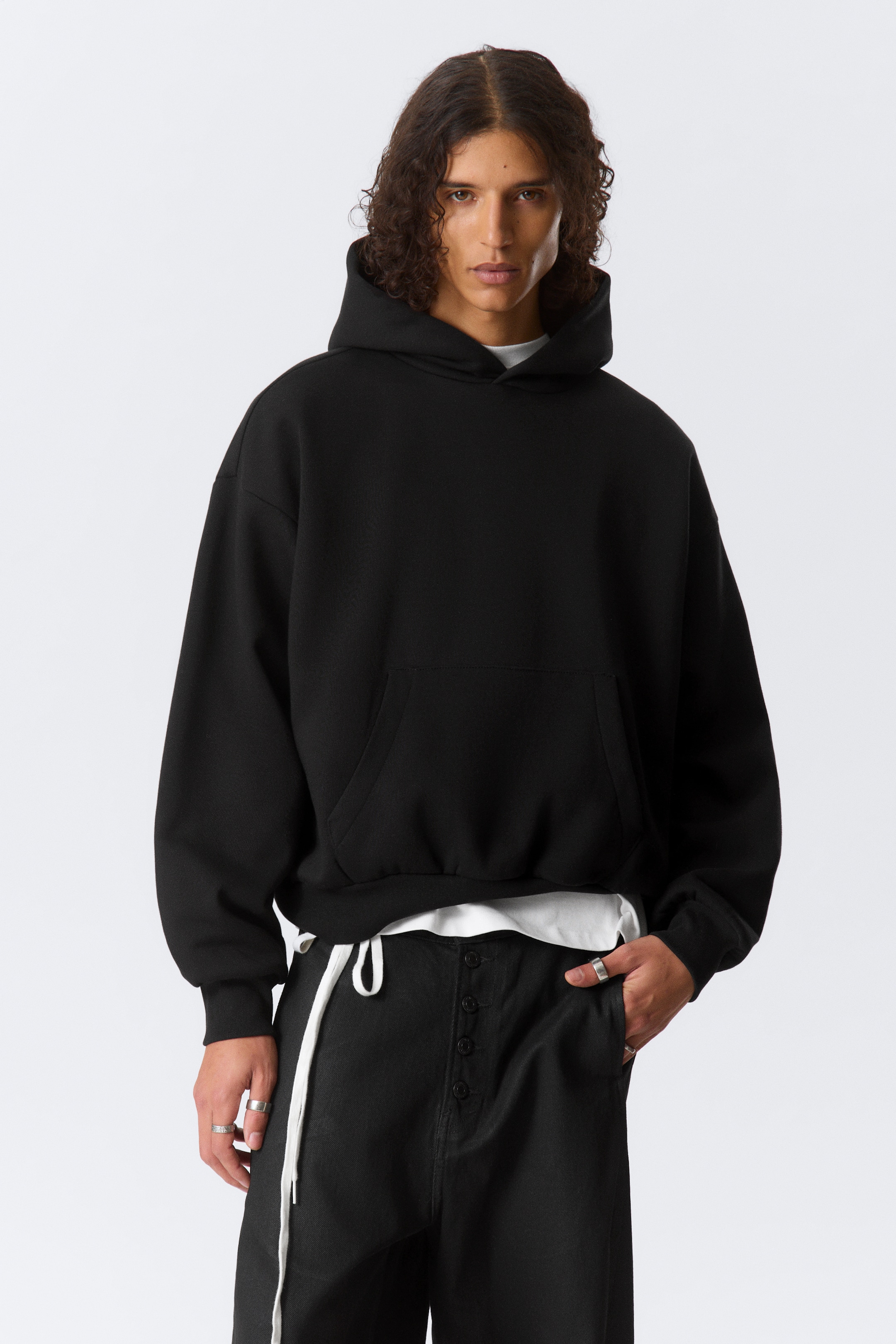 Grotere afbeelding bekijken: De zwarte oversized hoodie heeft een ontspannen pasvorm met verlaagde schouders en een grote kangoeroezak aan de voorkant. Het heeft een capuchon, geribbelde boorden en een geribbelde zoom, met een wit t-shirt dat zichtbaar onder de onderrand gelaagd is.
