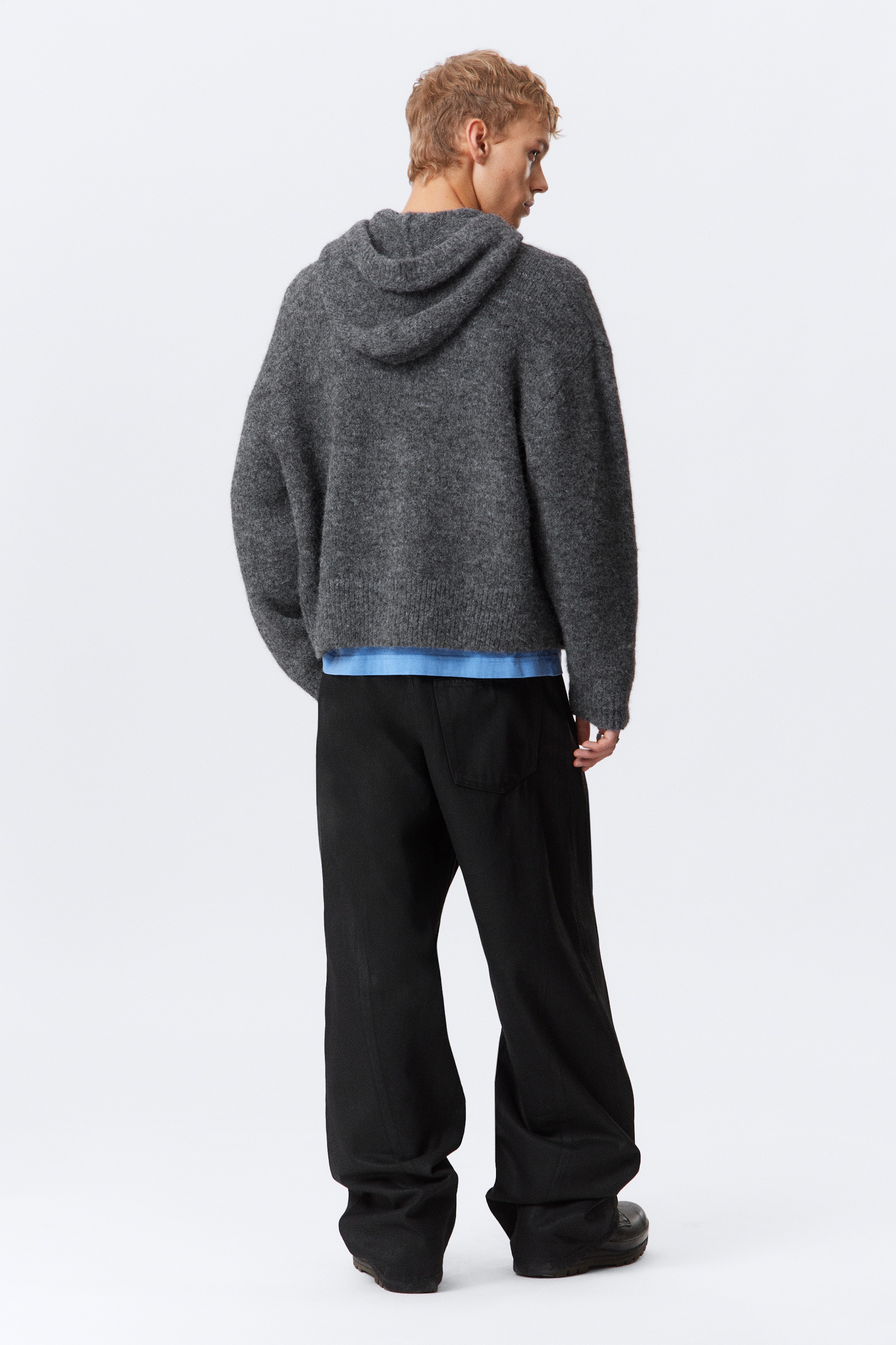View larger image: A dark grey knitted hoodie is being worn, viewed from the back. The oversized hoodie is featuring a hood and a ribbed hem, with a blue T-shirt visible peeking out from underneath the hem.