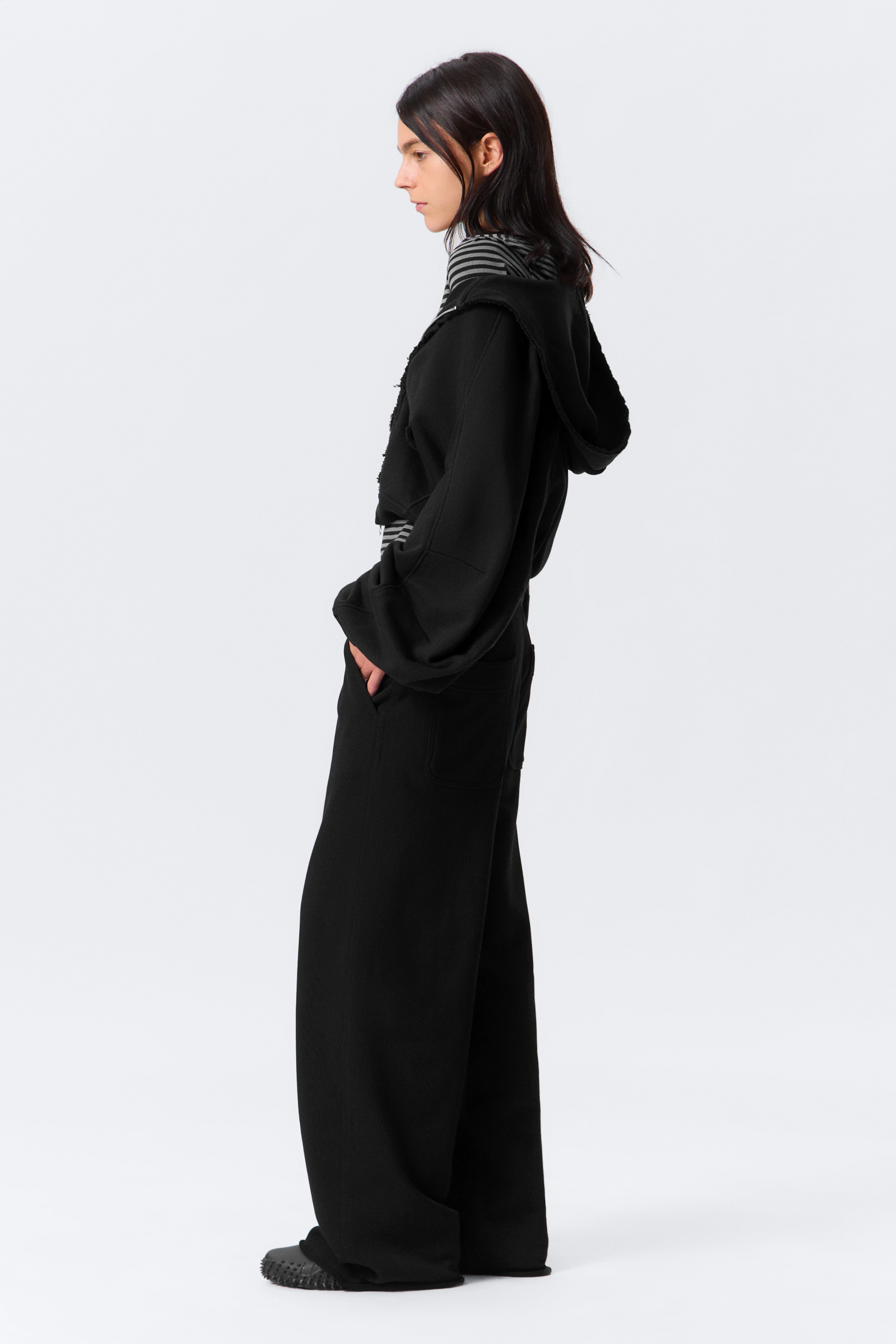 Visualizza immagine più grande: Pantaloni della tuta neri, larghi e ampi con gamba larga realizzati in un tessuto pesante sono indossati dalla modella. I pantaloni della tuta presentano orli non lavorati e tasche laterali inclinate, in una delle quali la mano della modella è appoggiata.