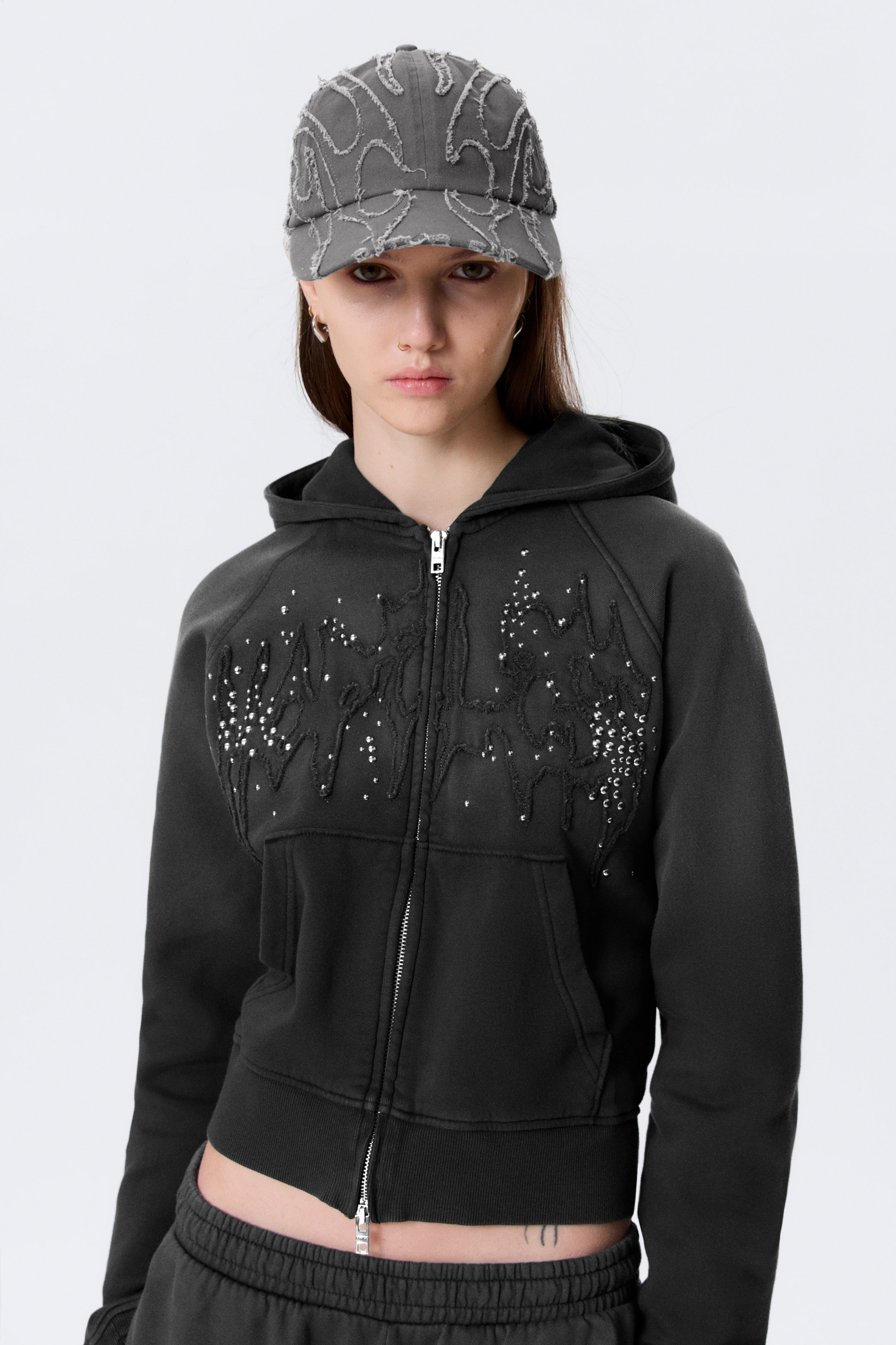 Grotere afbeelding bekijken: Een donkergrijze hoodie met volledige rits is naar voren gericht, met een verkorte lengte, geribbelde boorden en zoom, en kangoeroezakken. Een ingewikkeld distressed appliqué-ontwerp, versierd met talrijke zilveren studs, siert de voorkant van de borst.