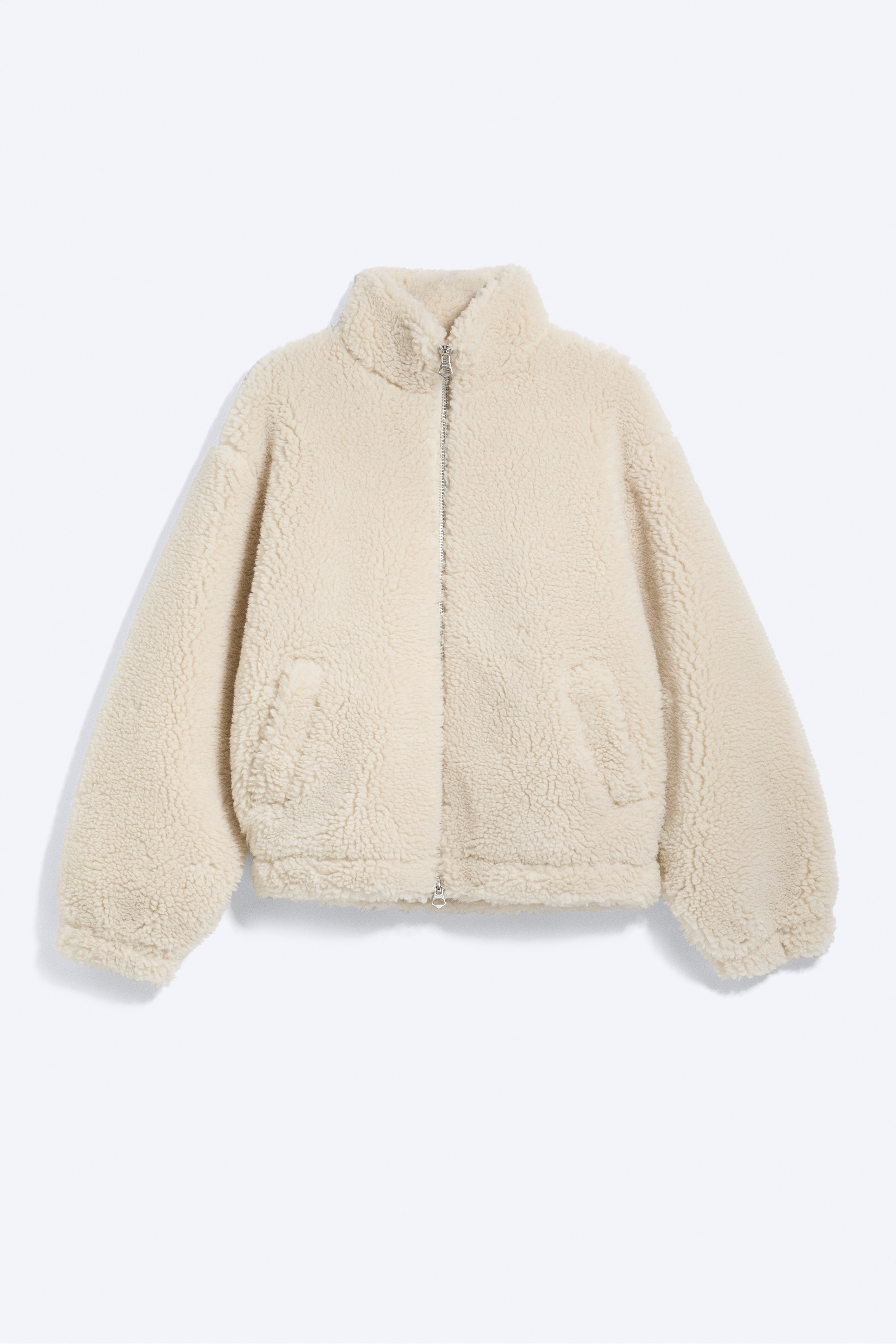 Una chaqueta de pelo color crema con cuello alto y silueta de corte holgado lleva puesta la modelo, que tiene una mano metida en el bolsillo delantero. La chaqueta se combina con un gorro de punto negro y pantalones de cuadros.