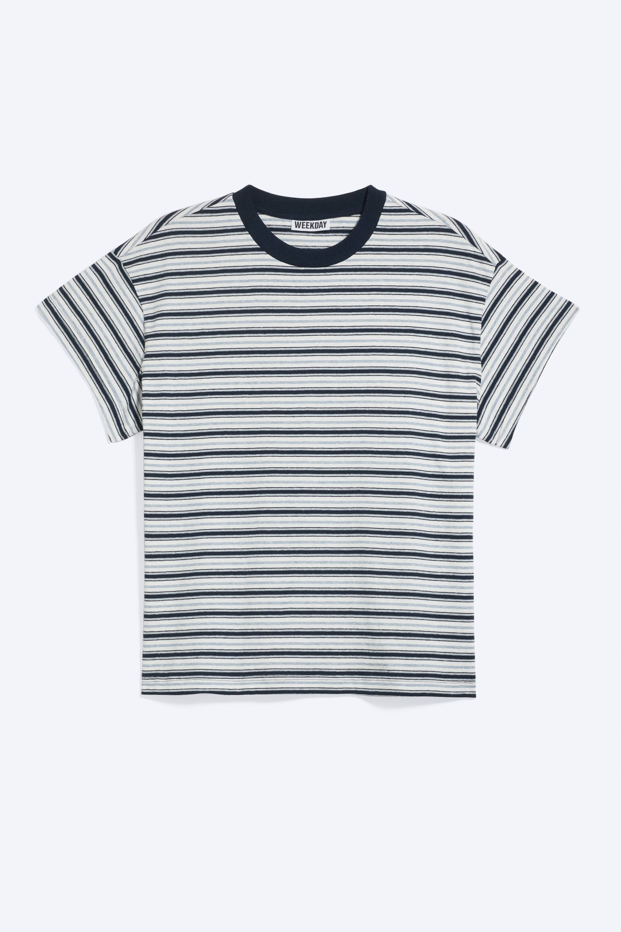 Agrandir l'image: Un T-shirt boxy est présenté de face, avec des rayures horizontales bleu foncé, bleu clair et blanc cassé. Il est conçu avec des épaules légèrement tombantes, des manches courtes à petites manches cap, ainsi que des détails usés au niveau de l’encolure côtelée et de l’ourlet, créant un effet vieilli.