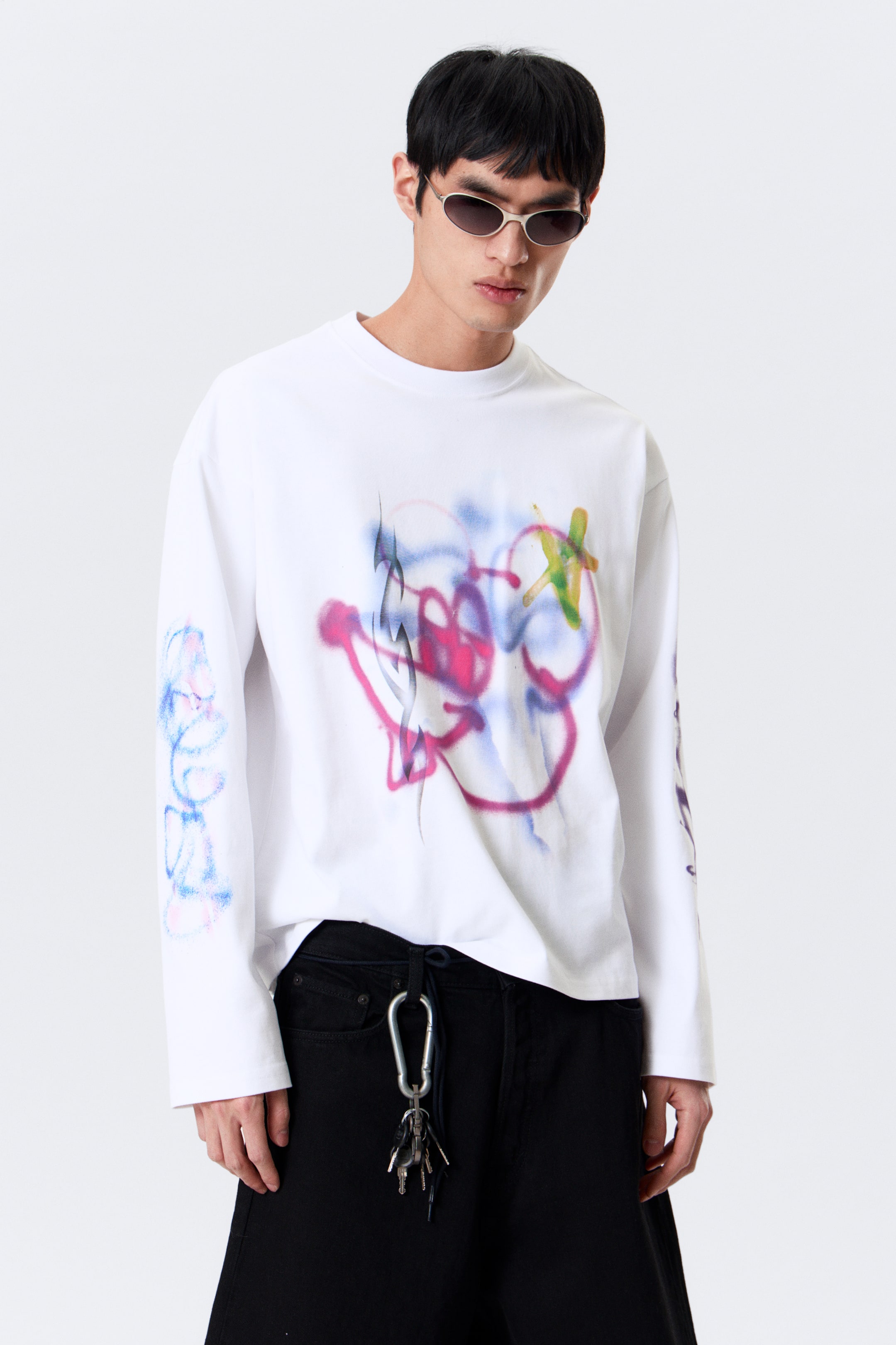 Zobraziť väčší obrázok: Biele tričko s dlhým rukávom zobrazuje viacfarebnú, sprejovo-maľovanú abstraktnú grafiku cez prednú časť hrudníka, ktorá obsahuje prekrývajúce sa ružové, fialové, modré, zelené a žlté znaky. Menšie, podobné čmáranice sa objavujú na vonkajších stranách oboch dlhých rukávov. Tričko je navrhnuté s okrúhlym výstrihom a jednoduchými manžetami, ponúka uvoľnený strih z predného pohľadu.