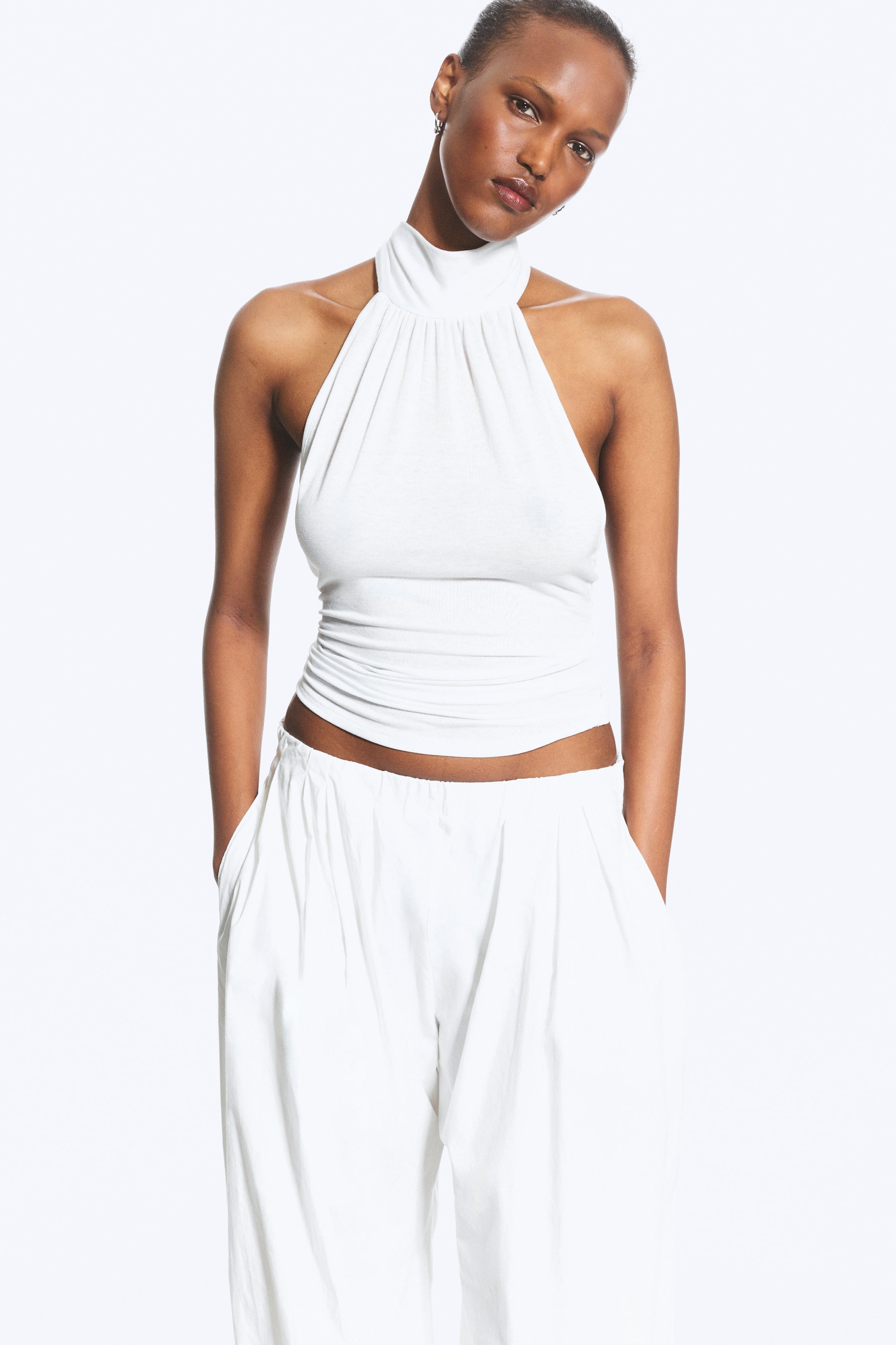 Größeres Bild ansehen: Ein weißes Neckholder-Top präsentiert einen hohen, gerafften Stehkragen und ein eng anliegendes, kurzes Oberteil. Es wird mit locker sitzenden, weißen Hosen mit weitem Bein kombiniert, die über einen elastischen Bund verfügen.