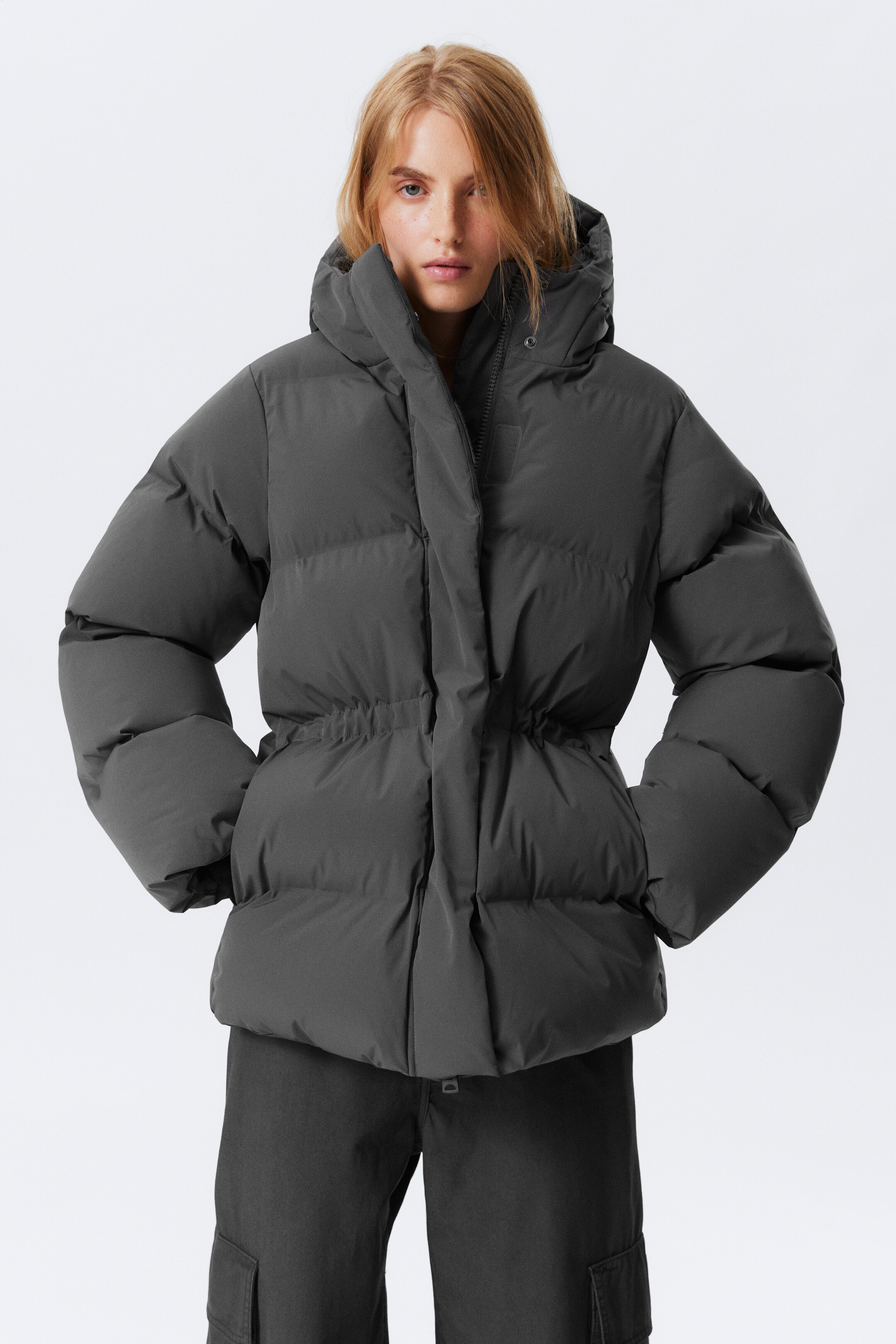 Größeres Bild anzeigen: Ein dunkelgrauer taillierter Steppjacke wird von dem Model getragen. Sie verfügt über eine Kapuze mit Kordelzug, eine vordere Druckknopfleiste und einen elastischen Kordelzug in der Taille, der die Jacke tailliert. Das Model steckt beide Hände in die Seitentaschen der Jacke, und unter der Jacke sind dunkle Hosen sichtbar.