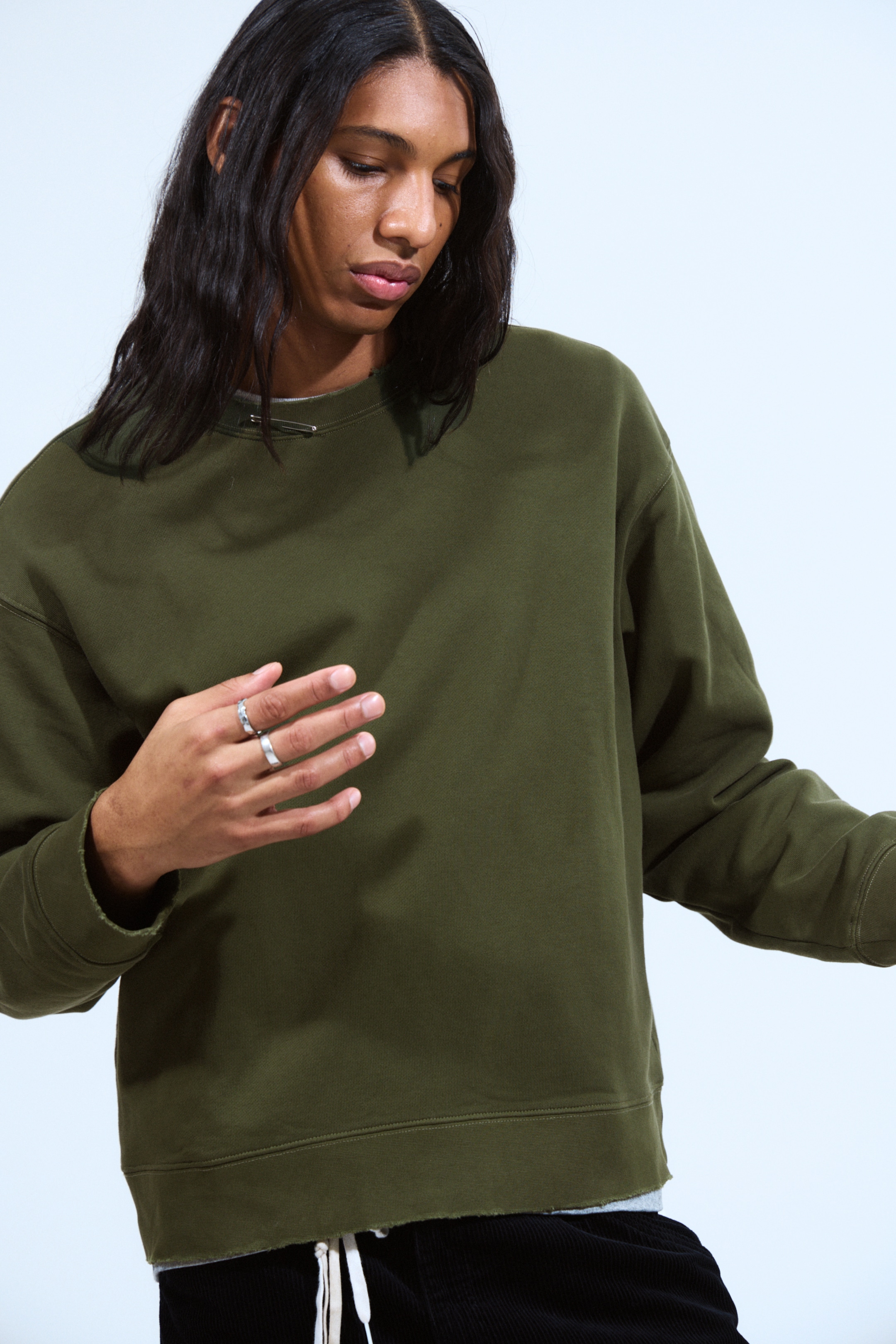 Agrandir l'image: Un sweat-shirt à col rond vert olive foncé est présenté sous un angle de 3/4 avant, avec une coupe décontractée, des épaules tombantes et un ourlet à bande avec un bord brut et usé, révélant un vêtement superposé plus clair en dessous. Une épingle de sûreté argentée est attachée au côté gauche du col rond.
