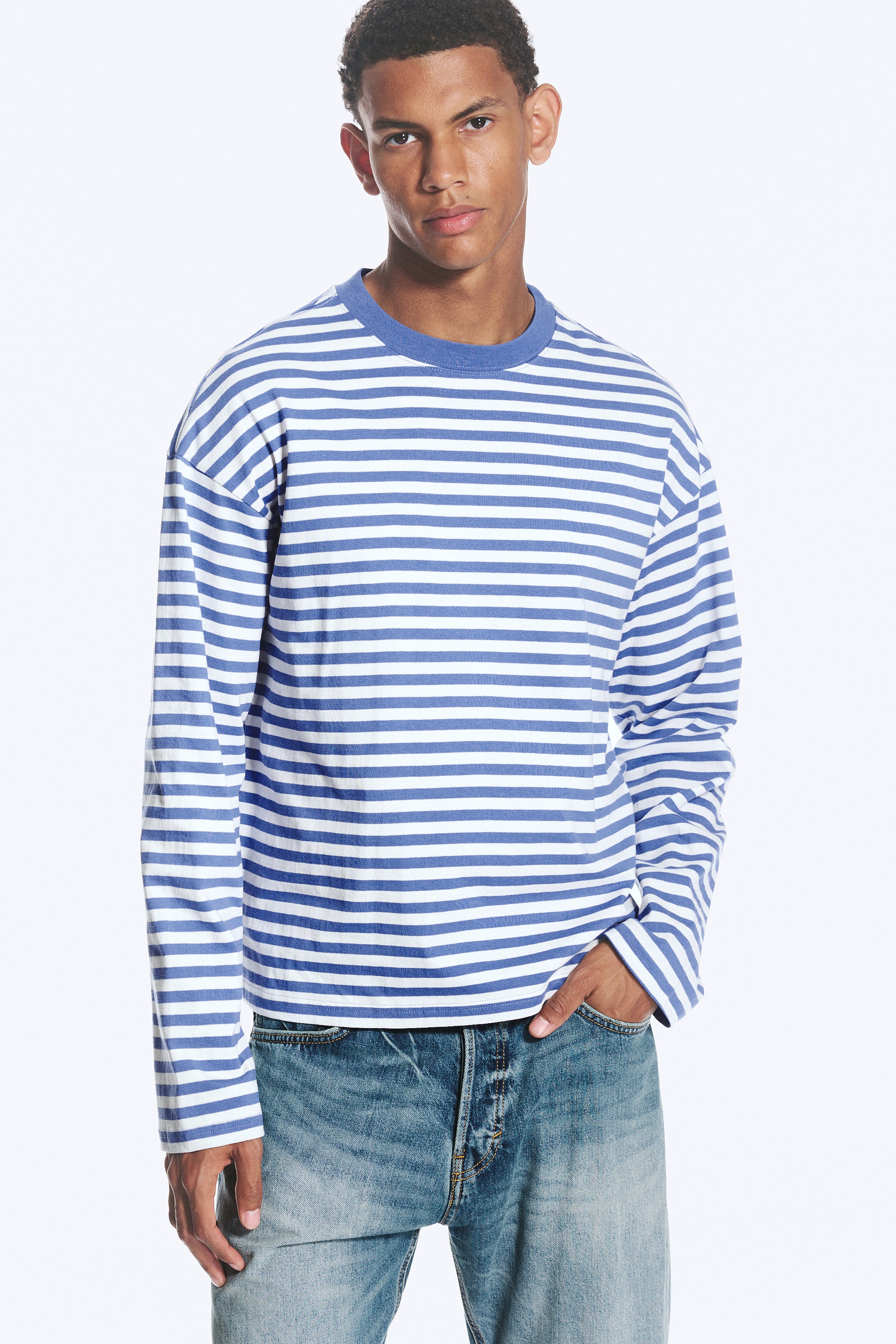 Ver imagem maior: A modelo está a usar uma t-shirt de manga comprida com riscas horizontais azuis e brancas. Apresenta uma gola redonda em malha canelada azul, um corte largo e bainhas direitas, com a mão da modelo a repousar num bolso de jeans azuis visíveis.