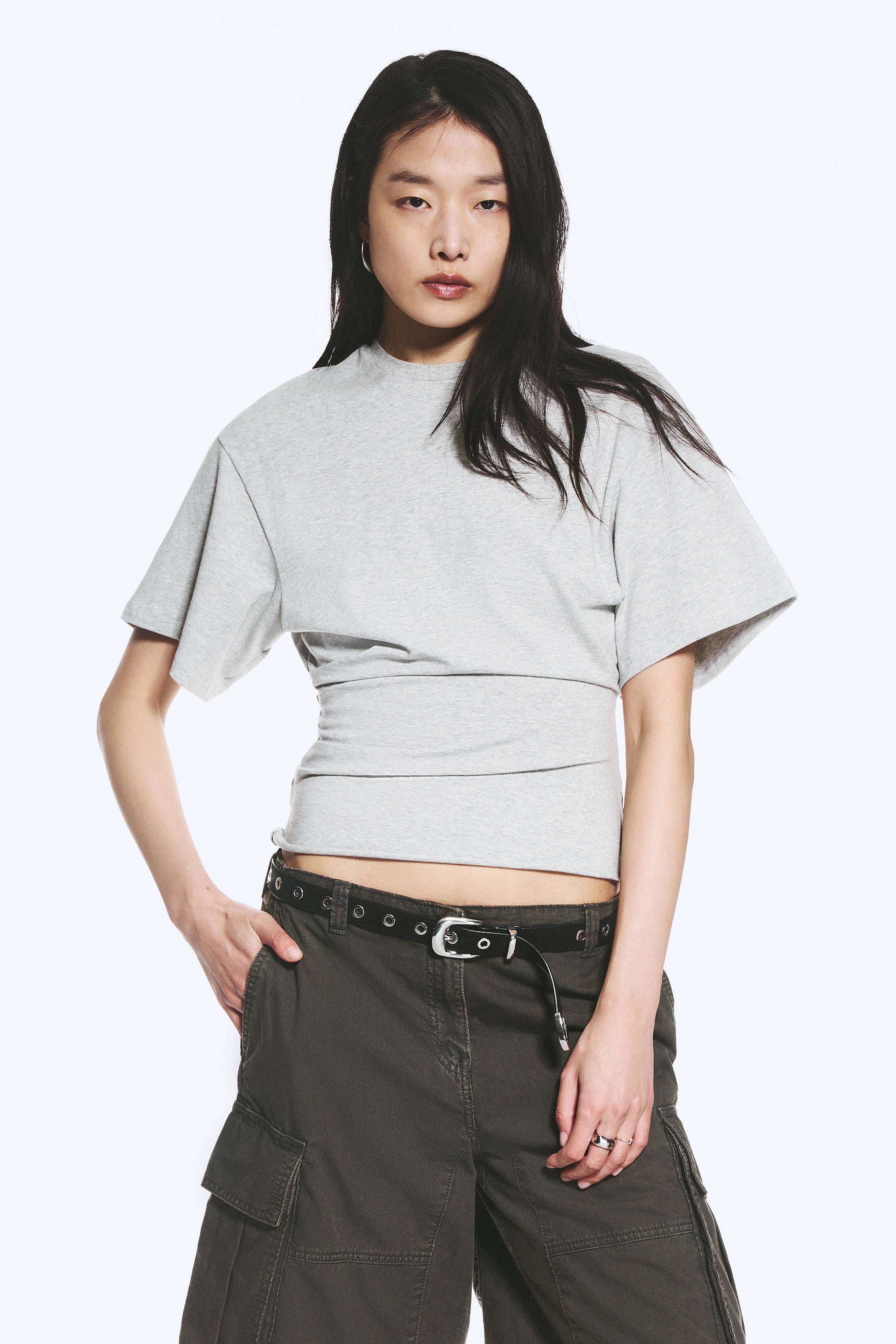 Se større billede: En lys grå tætsiddende T-shirt bæres af modellen, med boxy korte ærmer, rund halsudskæring og placerede læg. Disse læg skaber en draperet, tætsiddende silhuet omkring torsoen, der slutter over taljen, og T-shirten fremviser rå kantdetaljer.