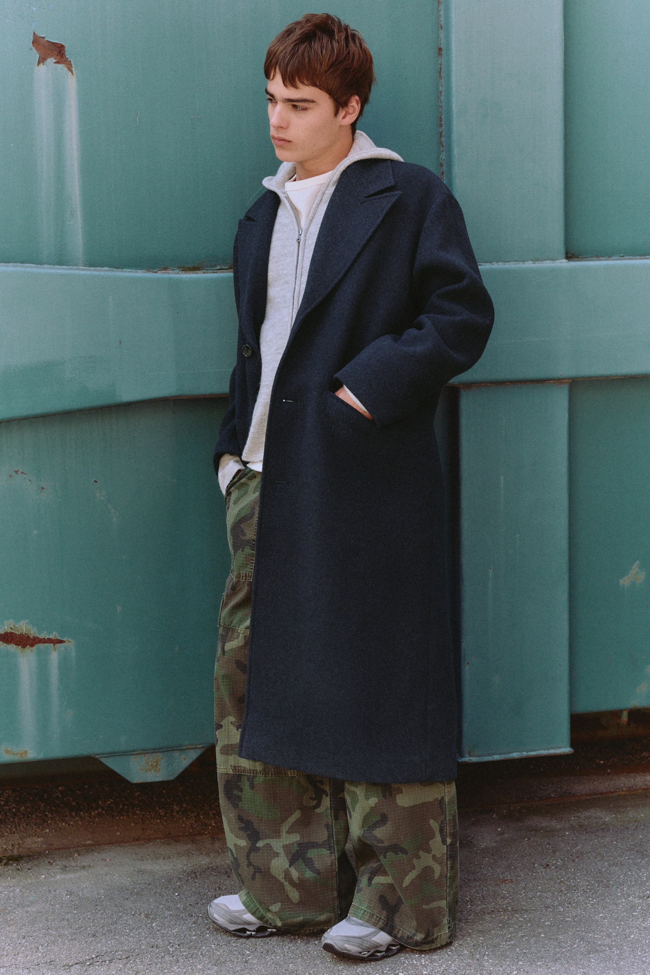 Zobrazit větší obrázek: Model má na sobě dlouhý oversized kabát v tmavě námořnické modré, stojí s jednou rukou zasunutou do boční kapsy. Kabát má klopový límec a uvolněný rovný střih, je stylizován přes světle šedou mikinu na zip a široké maskáčové kalhoty.