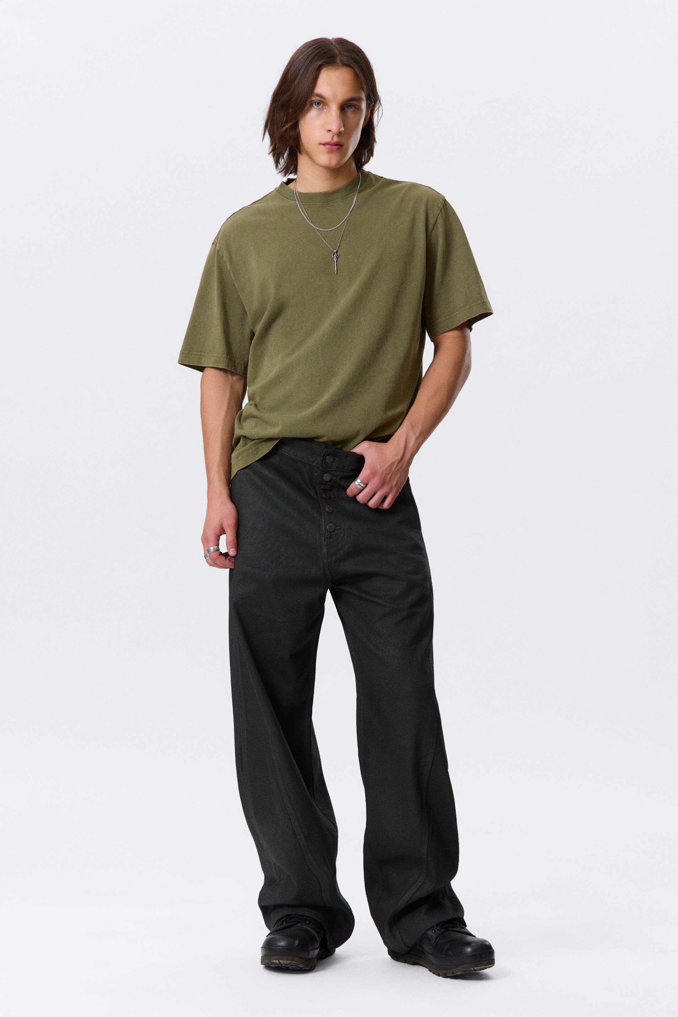 View larger image: An olive green, relaxed-fit t-shirt with a crew neck and short sleeves is being presented. It is being styled with dark gray, wide-leg pants featuring a prominent multi-button fly closure and a V-shaped waistband, the pants pooling loosely at the ankles.