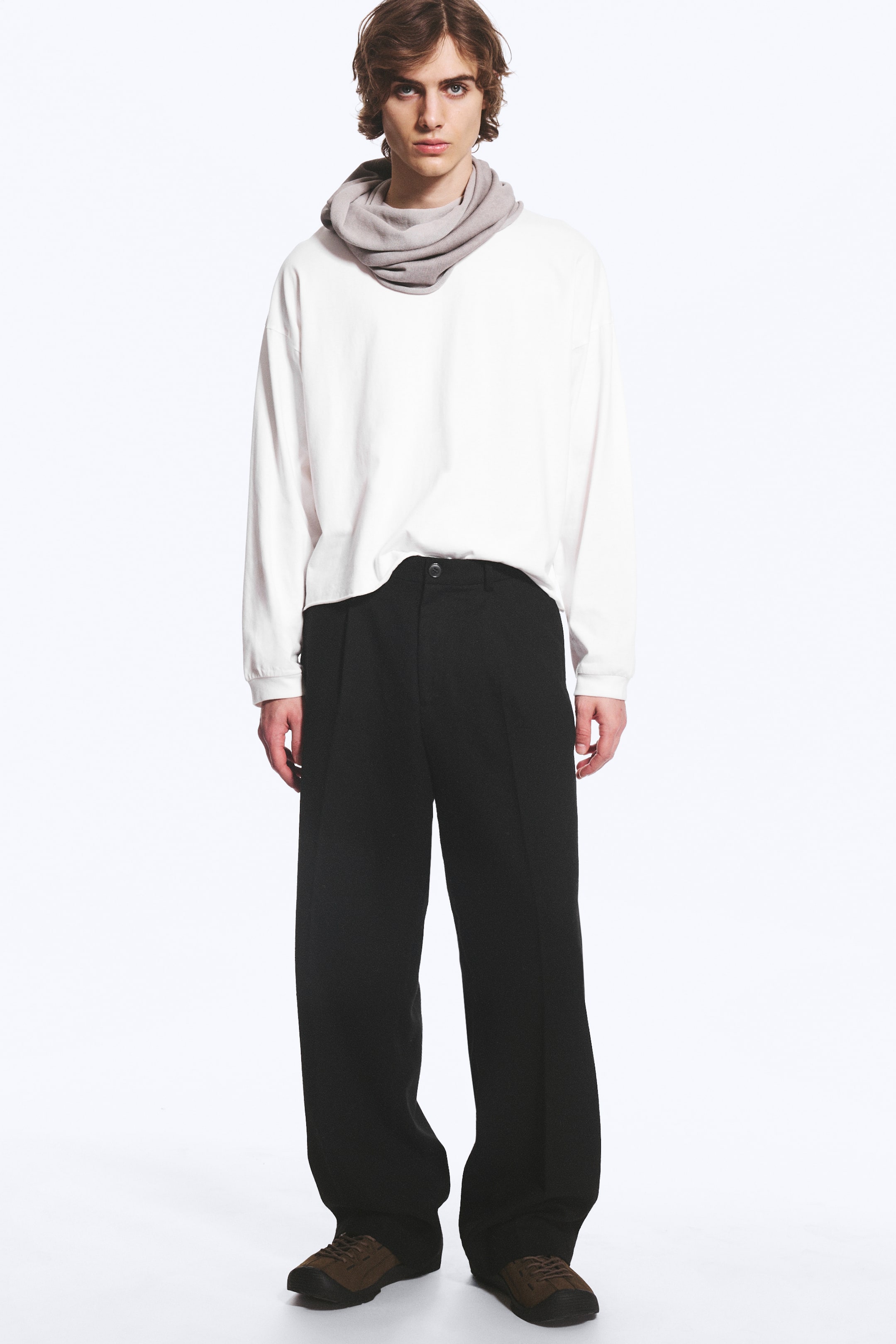 Visualizza l'immagine ingrandita: La modella indossa pantaloni neri a gamba ampia, caratterizzati da vita alta, pieghe frontali e una silhouette rilassata a gamba dritta. I pantaloni sono abbinati a una t-shirt bianca a maniche lunghe e a uno scaldacollo grigio drappeggiato.