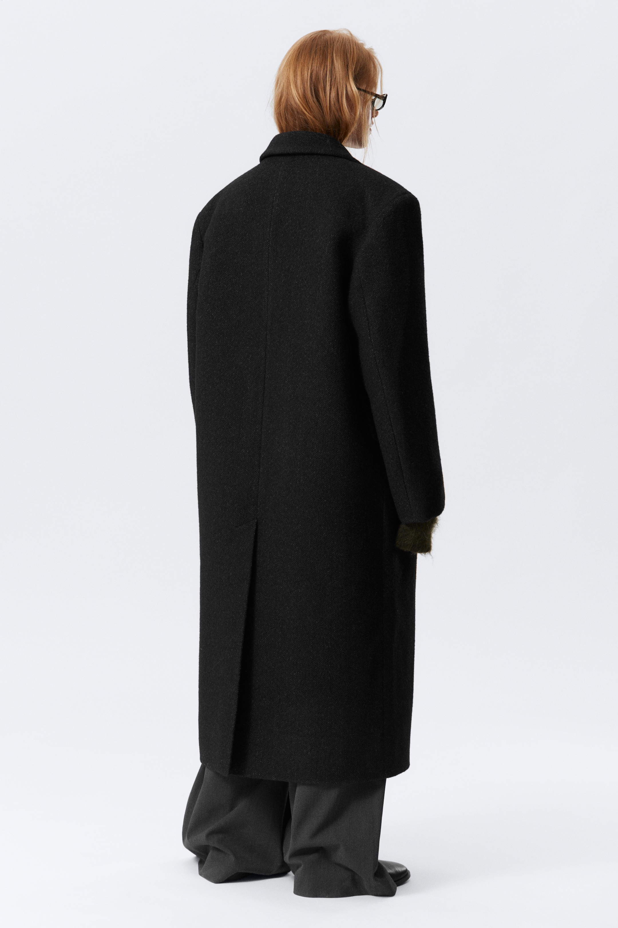 View larger image: A long, black coat is being viewed from the back, featuring a subtle textured fabric and a single back vent. It is being worn over a green garment, which is peeking out from the long sleeves, and dark grey wide-leg trousers are visible below the coat's hem.