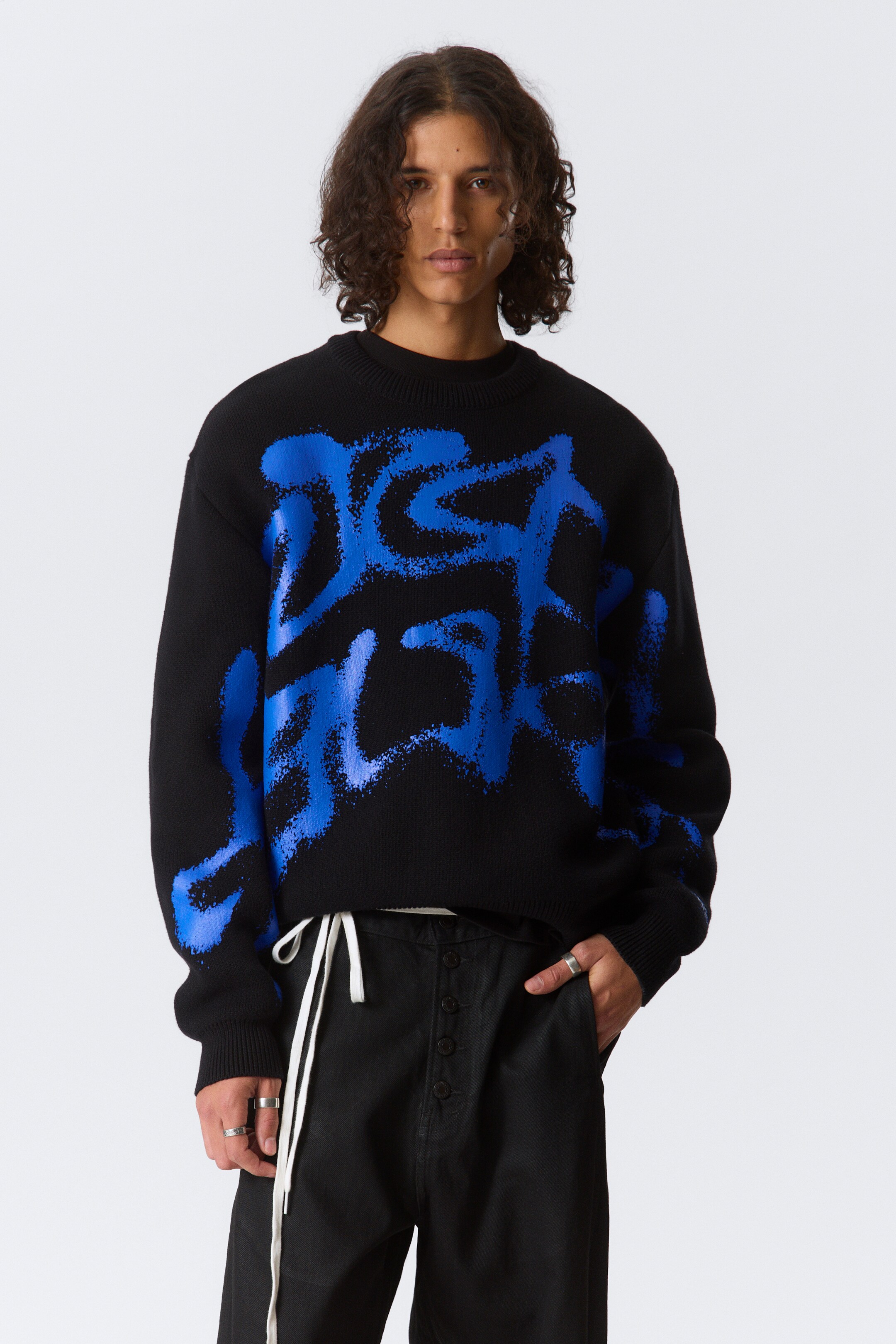 Se større billede: En sort crew-neck sweater har en stor, abstrakt blå grafik med en spraymalingseffekt på forsiden. Sweateren har ribbede manchetter og kant og præsenterer en afslappet pasform.