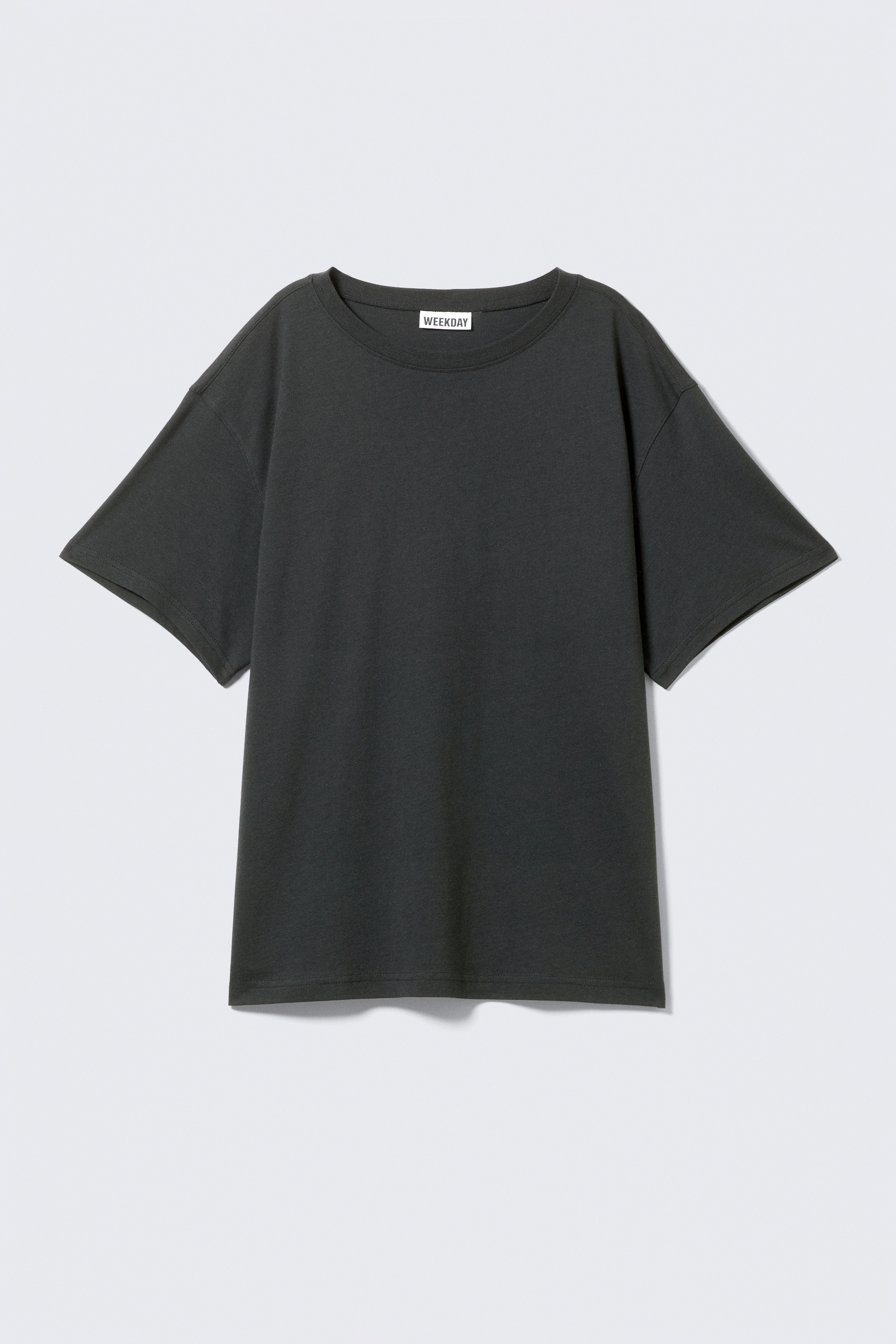 Vezi imaginea mai mare: Este prezentat un tricou gri închis, oversized, cu mâneci scurte, orientat spre FAȚĂ, având o siluetă relaxată cu umeri căzuți, un decolteu clasic rotund și o lungime mai mare, afișând o drapare subtilă. O etichetă albă de brand 'WEEKDAY' este vizibilă pe partea interioară a decolteului.