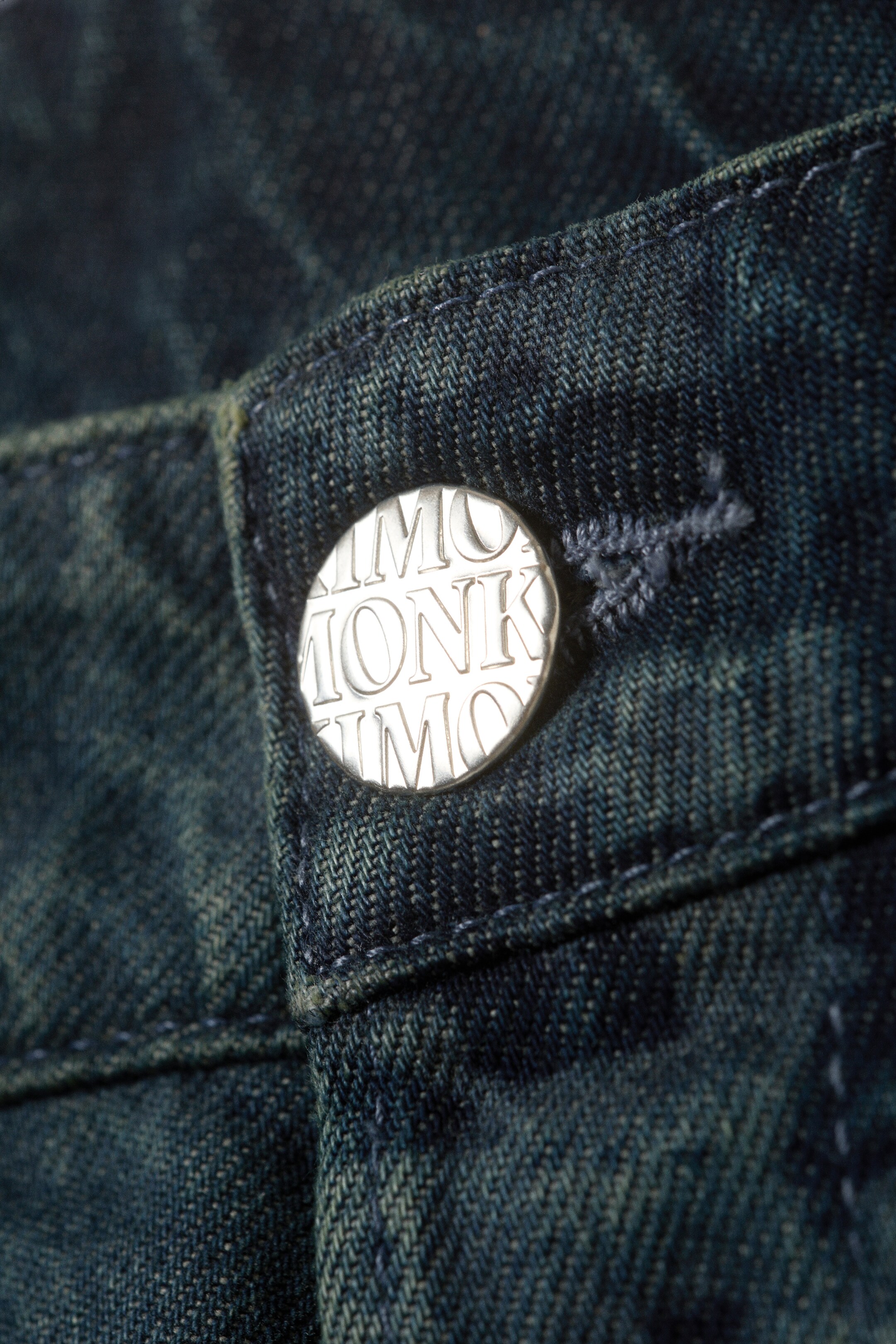 View larger image: Dark blue denim jeans are featuring a close-up view of their waistband, where a silver-toned button with embossed text is securing the front closure. The rigid denim fabric is displaying a textured weave and subtle wash effects.