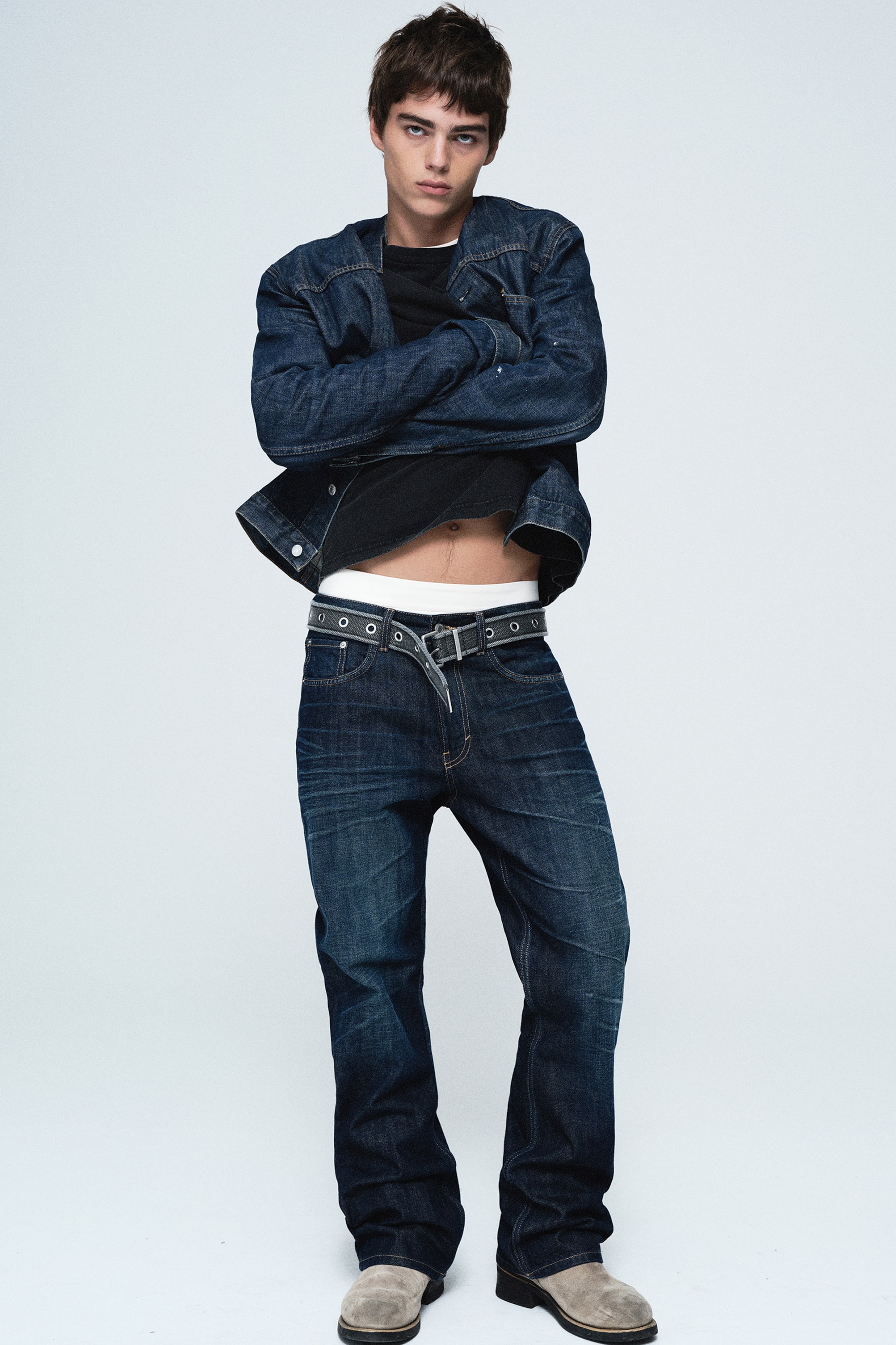 Agrandir l'image: Un jean en denim délavé foncé à coupe décontractée met en valeur une silhouette à jambe droite, avec un léger effet de moustaches au niveau des cuisses, vu de face. Il est porté avec une ceinture noire à œillets, portée bas pour laisser apparaître une ceinture de taille blanche, et associé à une veste en denim délavé foncé assortie, portée sur un t-shirt noir.