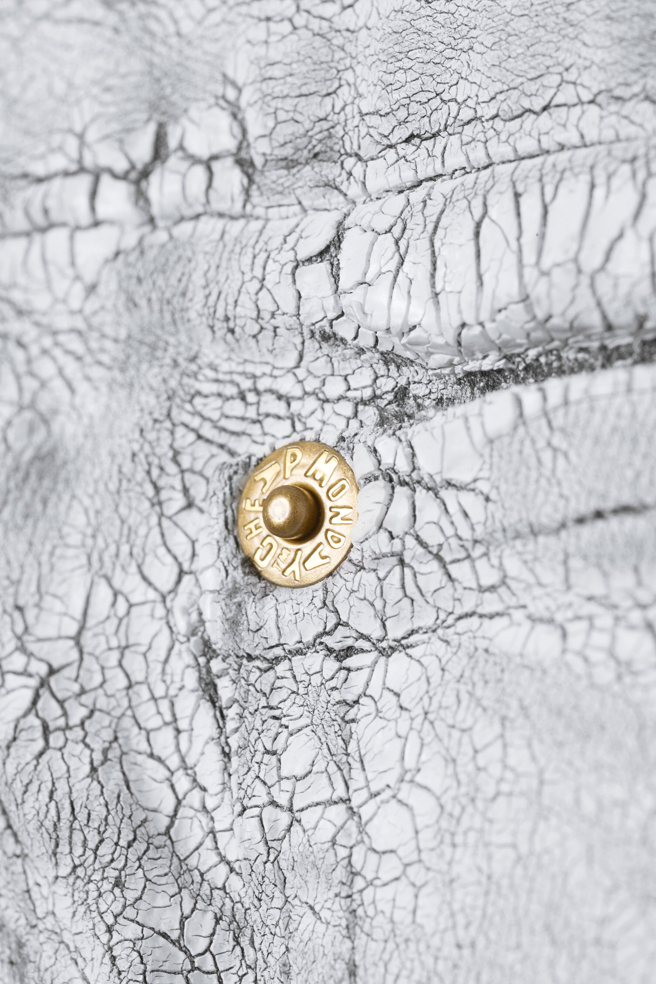 Agrandir l'image: Un matériau blanc avec une texture fortement craquelée affiche des lignes sombres dans ses crevasses. Un rivet circulaire de couleur laiton avec un centre surélevé et un texte en relief est visible sur le matériau.