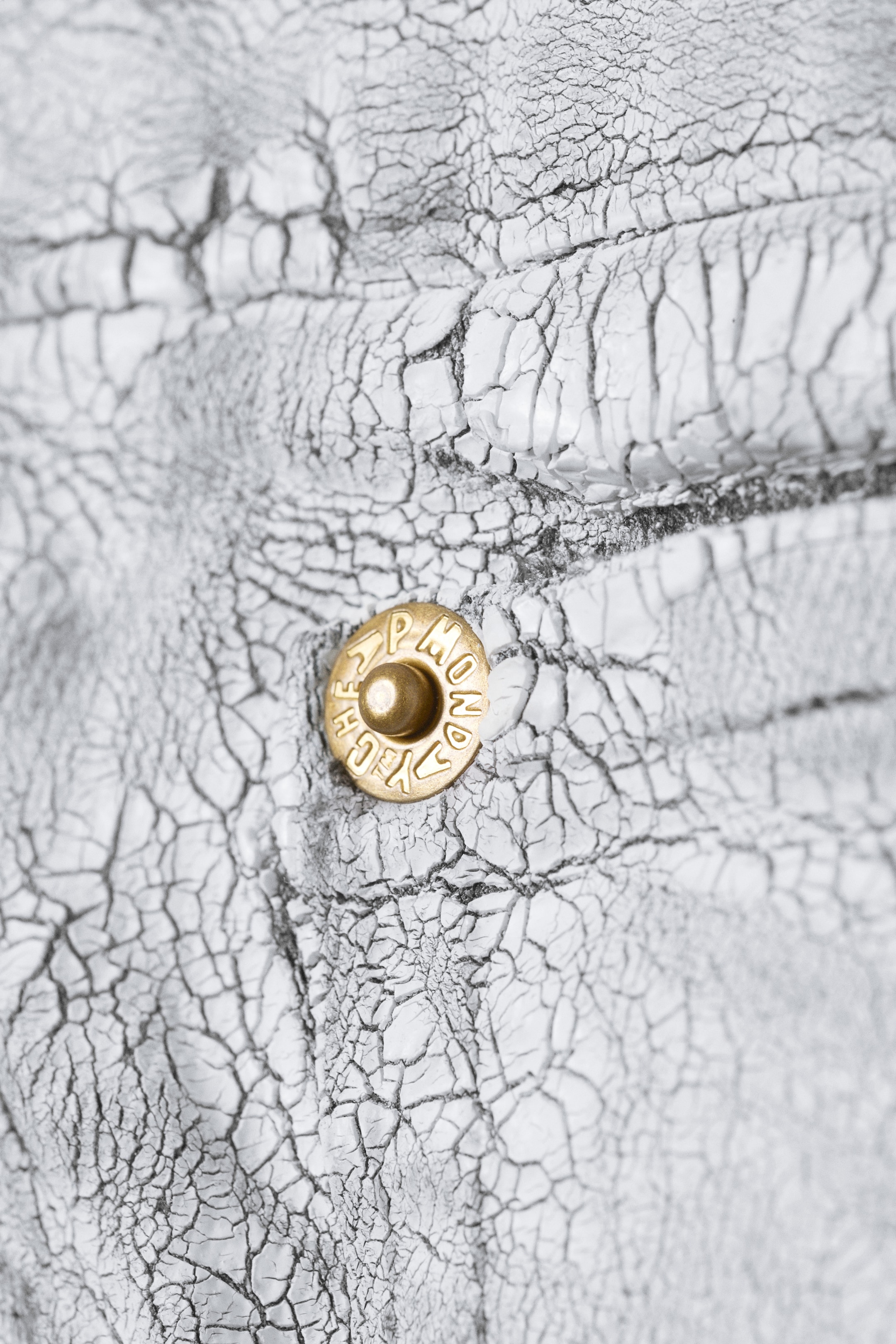 Agrandir l'image: Un matériau blanc avec une texture fortement craquelée affiche des lignes sombres dans ses crevasses. Un rivet circulaire de couleur laiton avec un centre surélevé et un texte en relief est visible sur le matériau.