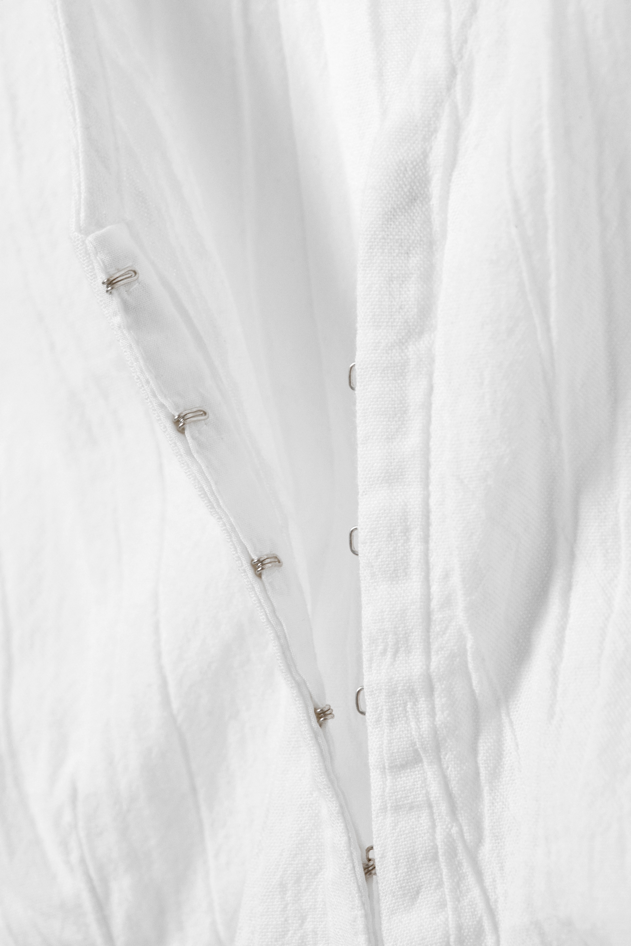 Visualizza immagine più grande: Un top bianco senza maniche presenta una texture stropicciata e una chiusura frontale dettagliata con multipli fermagli a occhiello in tono argento.