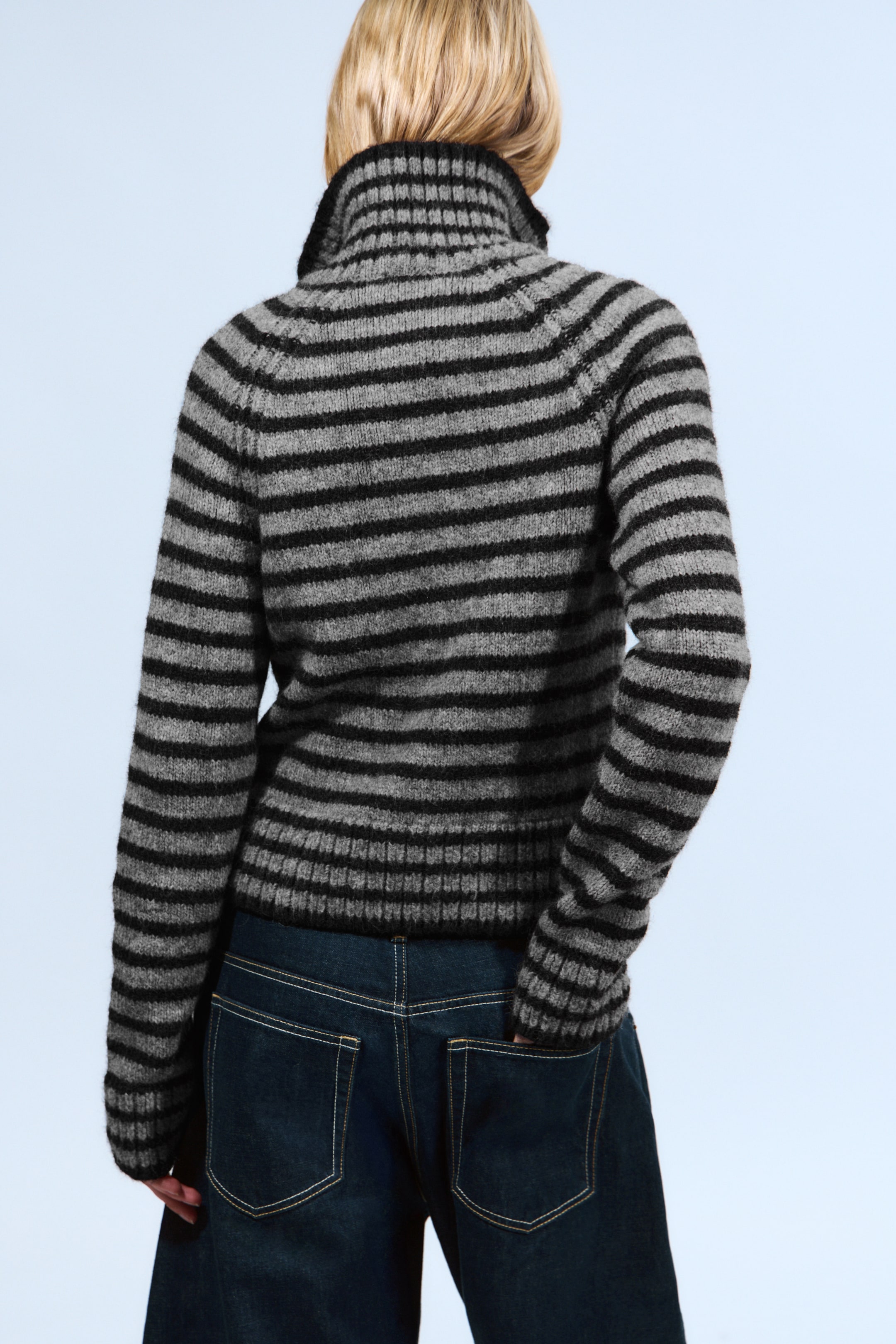 Visualizza immagine più grande: Un maglione a righe grigie e nere viene indossato dal retro, mostrando la sua trama lavorata a maglia e le righe orizzontali grigie e nere. Un colletto a collo alto a coste si alza, e le maniche raglan si estendono verso il basso, mentre un orlo a coste si appoggia sulla linea della vita.