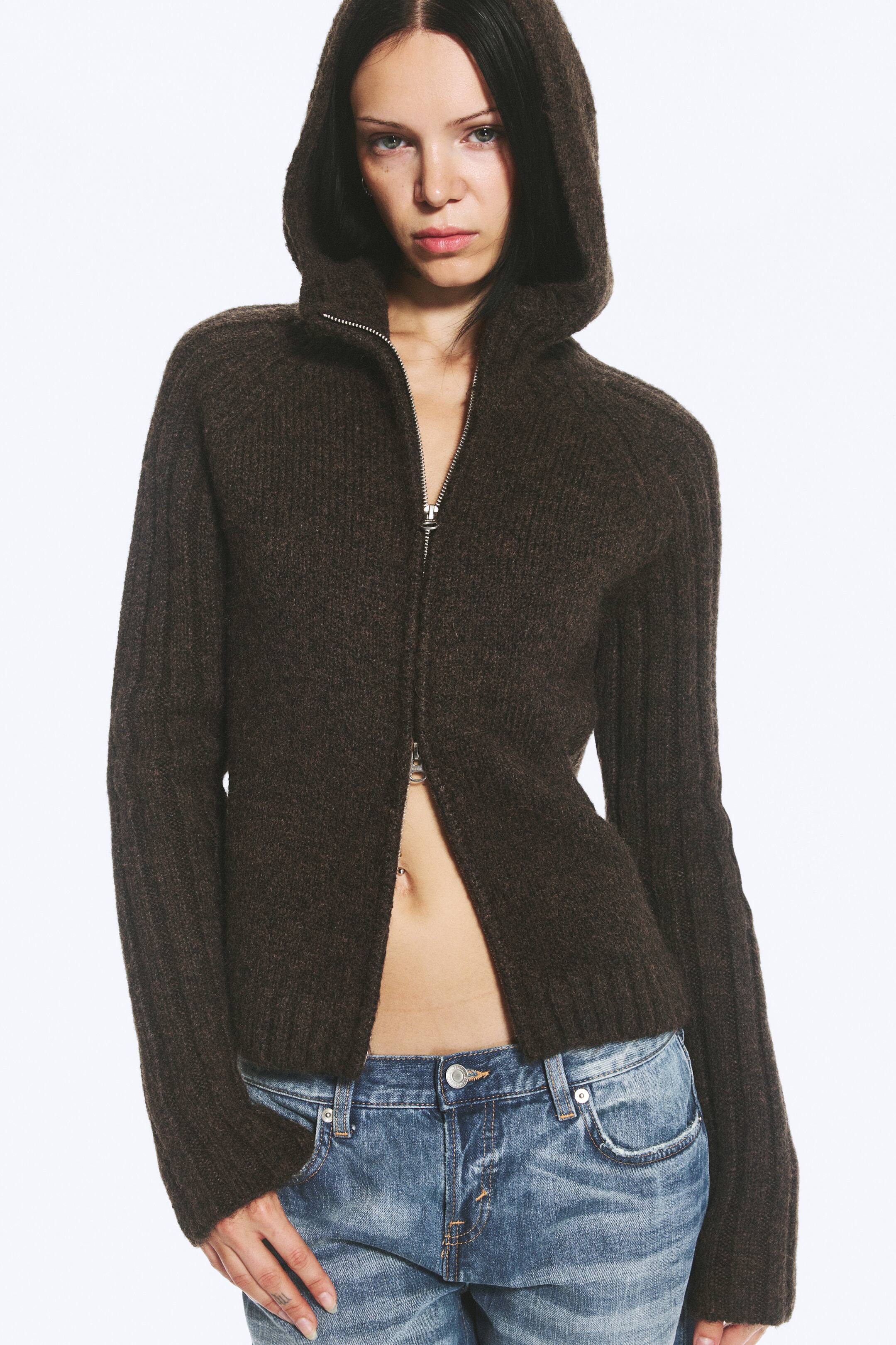 Ver imagen más grande: La modelo lleva puesta una sudadera con capucha de punto acanalado en color marrón oscuro, con la capucha subida. La sudadera presenta una cremallera frontal de doble sentido, parcialmente abierta por la parte superior e inferior, y se combina con unos vaqueros azules de tiro bajo.