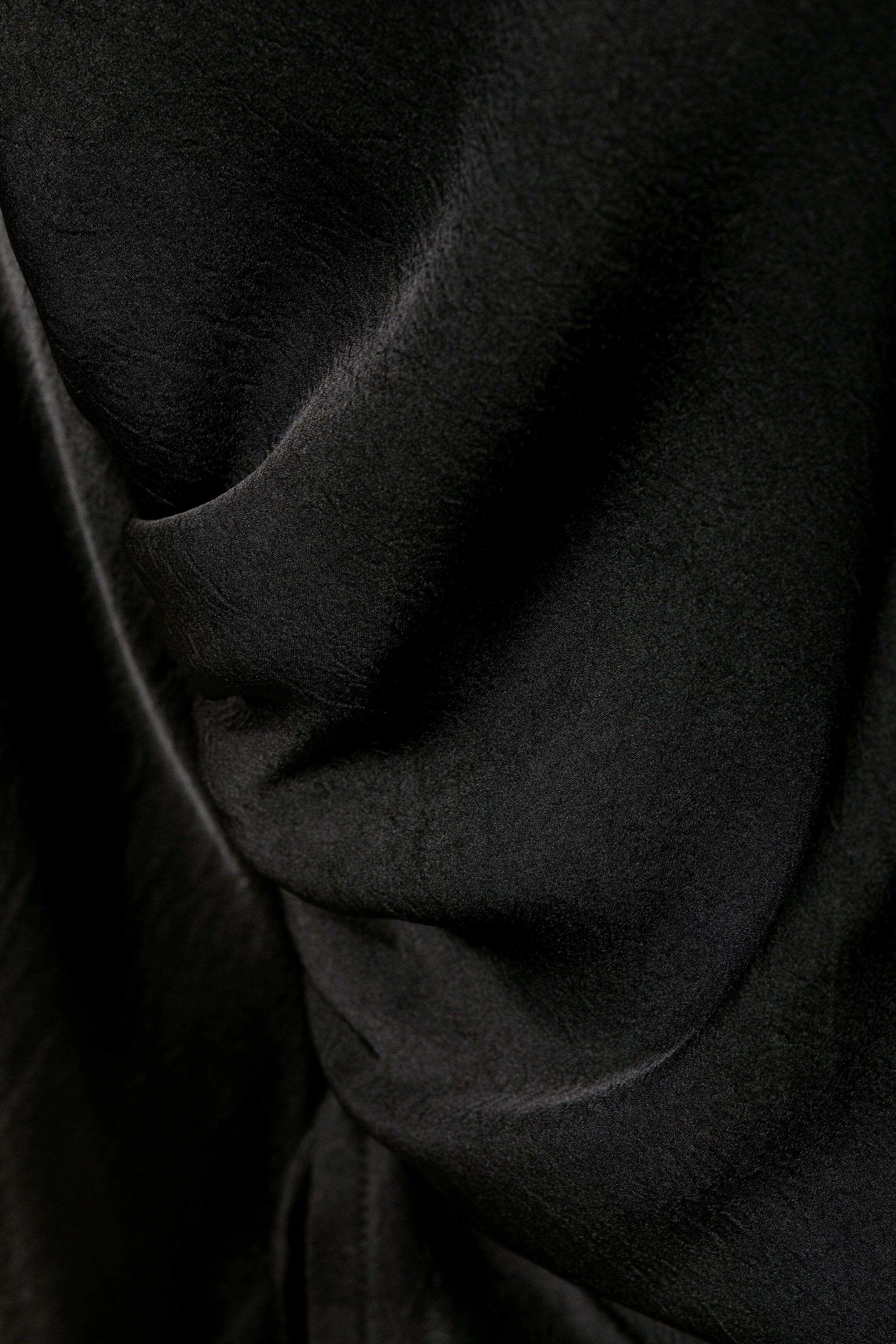 View larger image: A black fabric is displaying soft folds and a subtle crinkled texture. Light is catching on the raised areas of the material, creating variations in tone across its surface.