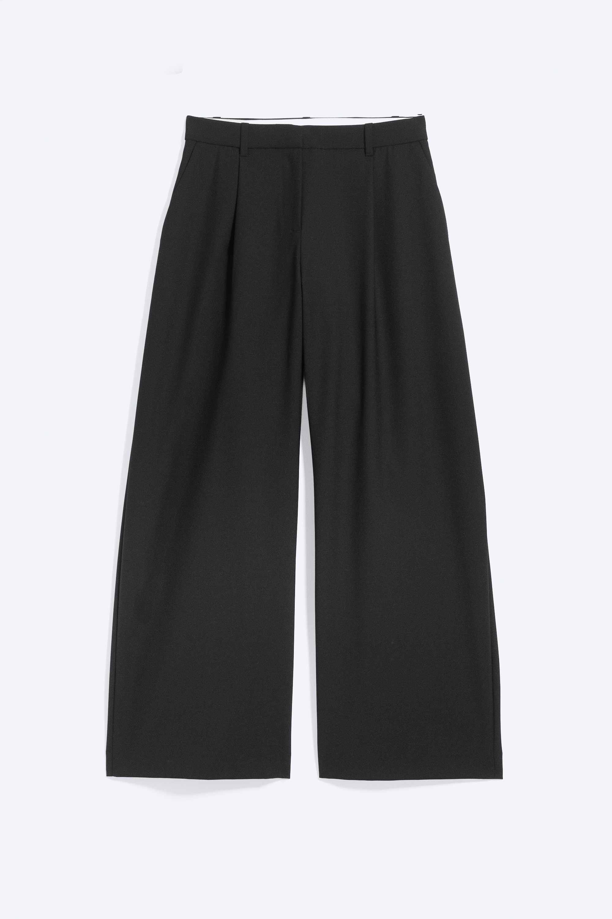 The model is wearing black, wide-leg tailored trousers, styled with a black belt threaded through the belt loops. She is lifting the hem of a dark, long-sleeved top to reveal the low-rise waist of the trousers.