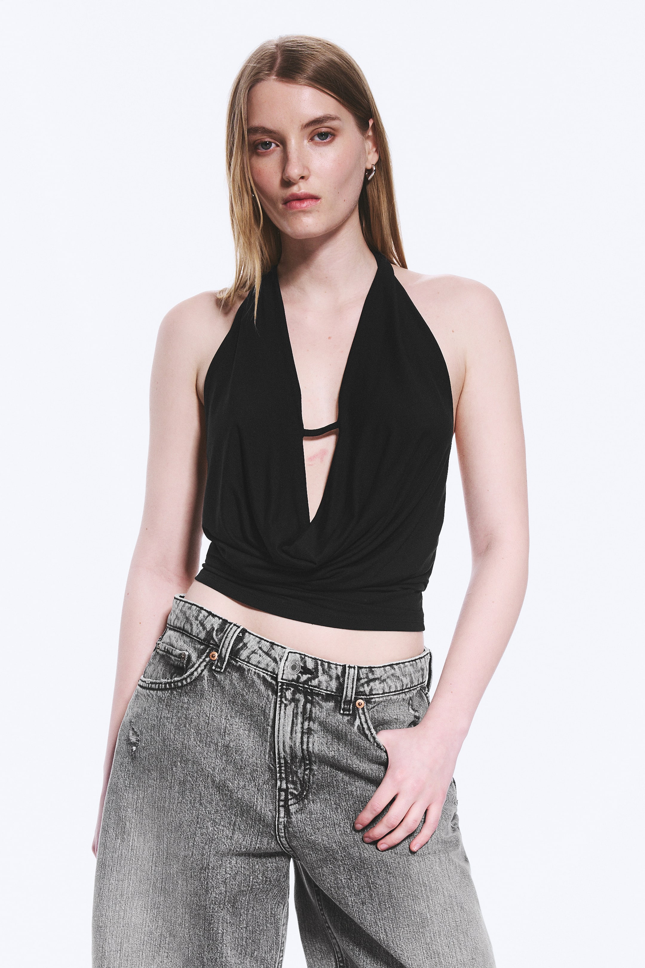 Größeres Bild ansehen: Ein schwarzes Neckholder-Top mit einem tiefen Wasserfallausschnitt und einer verkürzten Länge. Ein dünner schwarzer Riemen ist quer über der Brust sichtbar, unter dem drapierten Stoff des Ausschnitts.