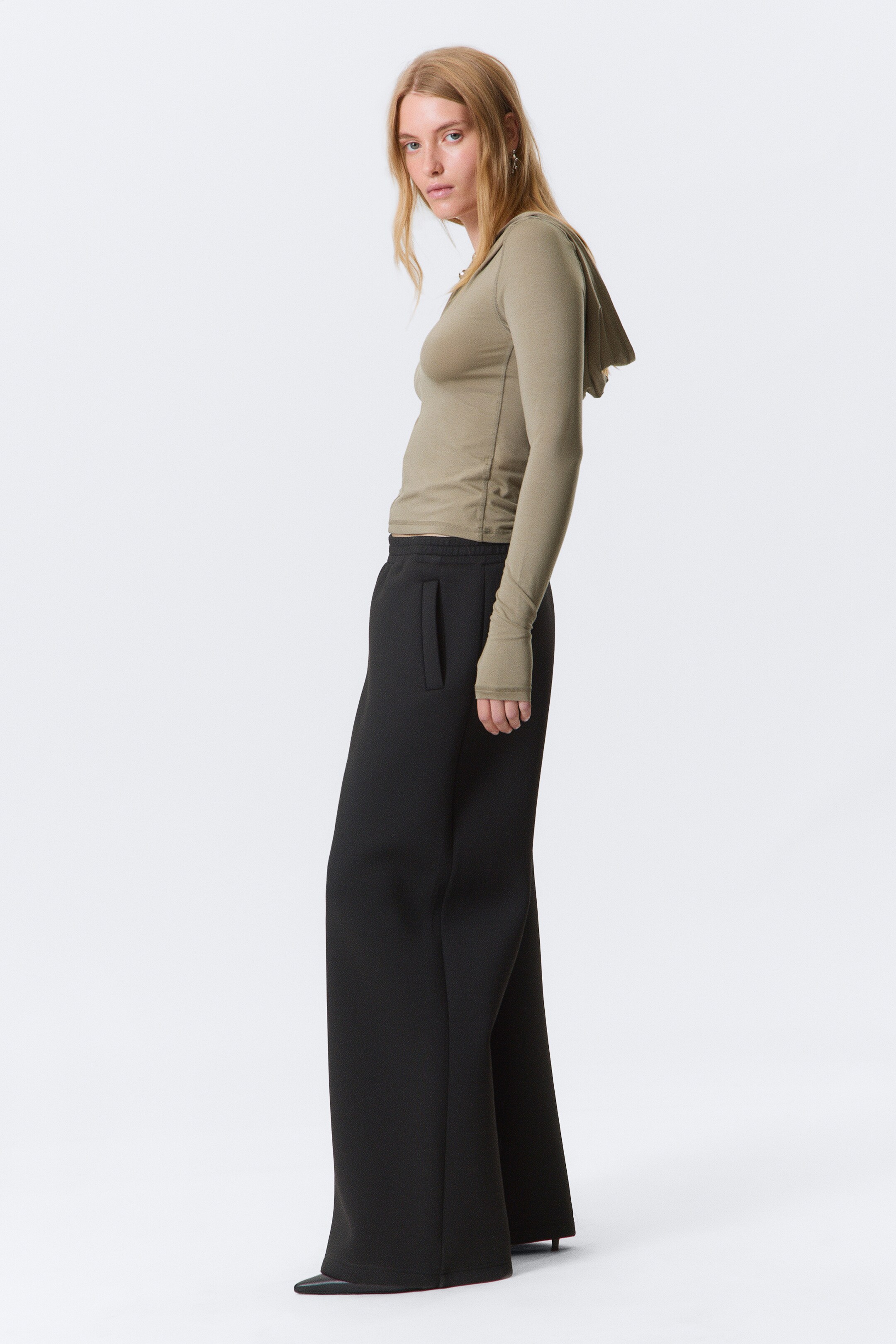 Ver imagen más grande: Pantalones de chándal negros de pierna ancha con una silueta estructurada están siendo usados por una modelo. Los pantalones cuentan con un solo bolsillo de ojal en la cadera derecha y una raya planchada en la parte delantera de cada pierna, combinados con un top ajustado de manga larga con capucha.