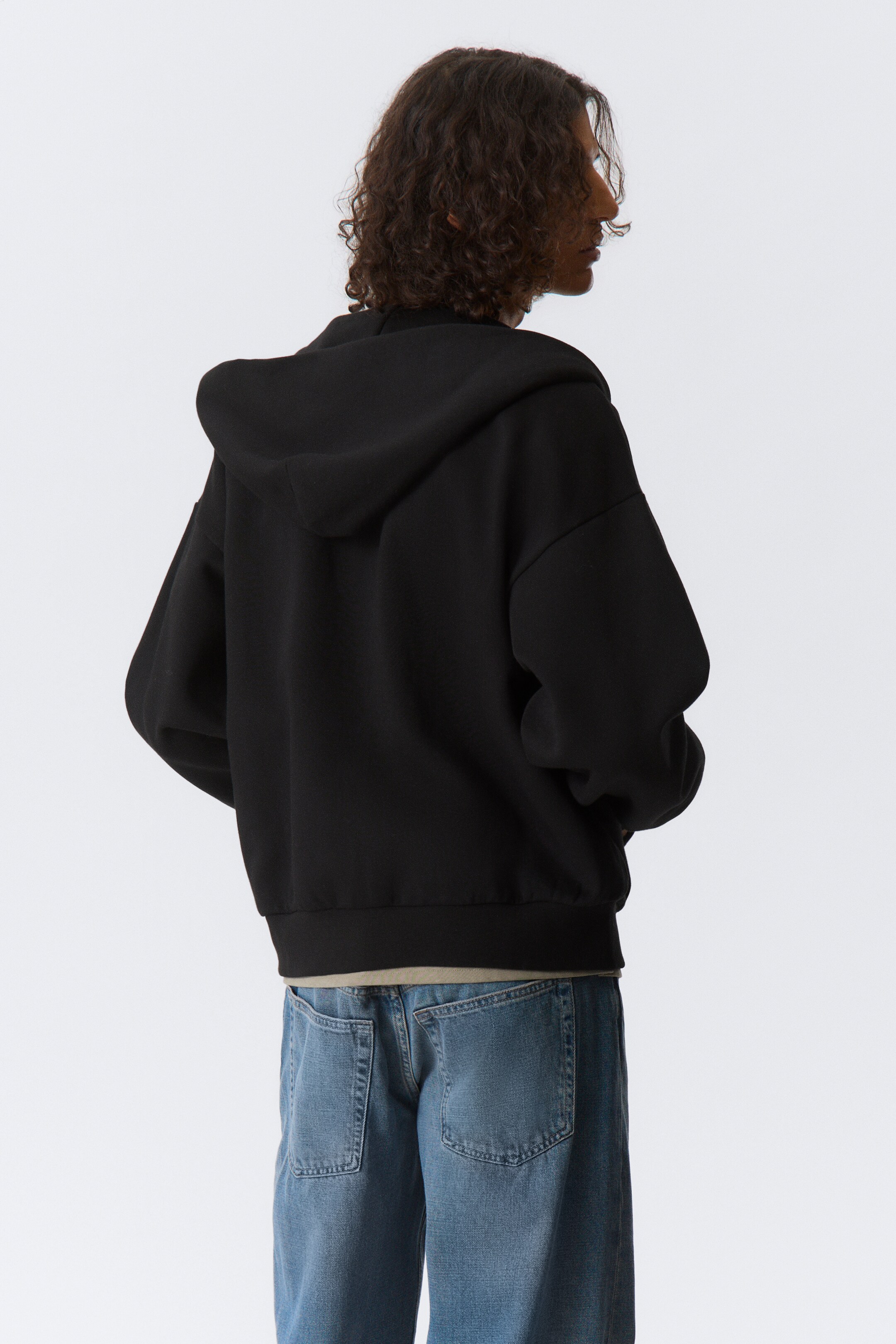Ver imagem maior: Uma hoodie preta é apresentada de trás para a direita, com corte descontraído, capuz, mangas compridas e bainha canelada, sobrepondo uma t-shirt de cor clara e jeans azuis.