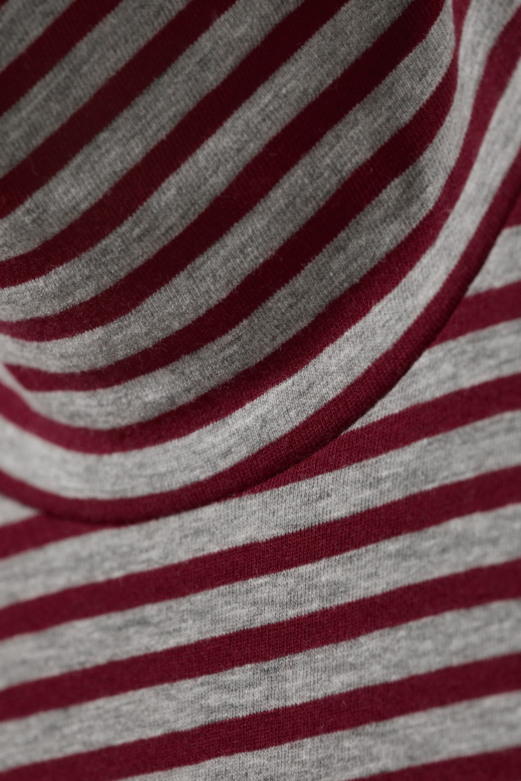 Größeres Bild anzeigen: Ein Rollkragenoberteil präsentiert sein grau-burgundfarbenes, horizontal gestreiftes Muster auf einem Jersey-Strickstoff. Eine gebogene, gefaltete Kante, die auf den doppellagigen Kragen hinweist, ist ebenfalls im oberen Rahmen sichtbar.