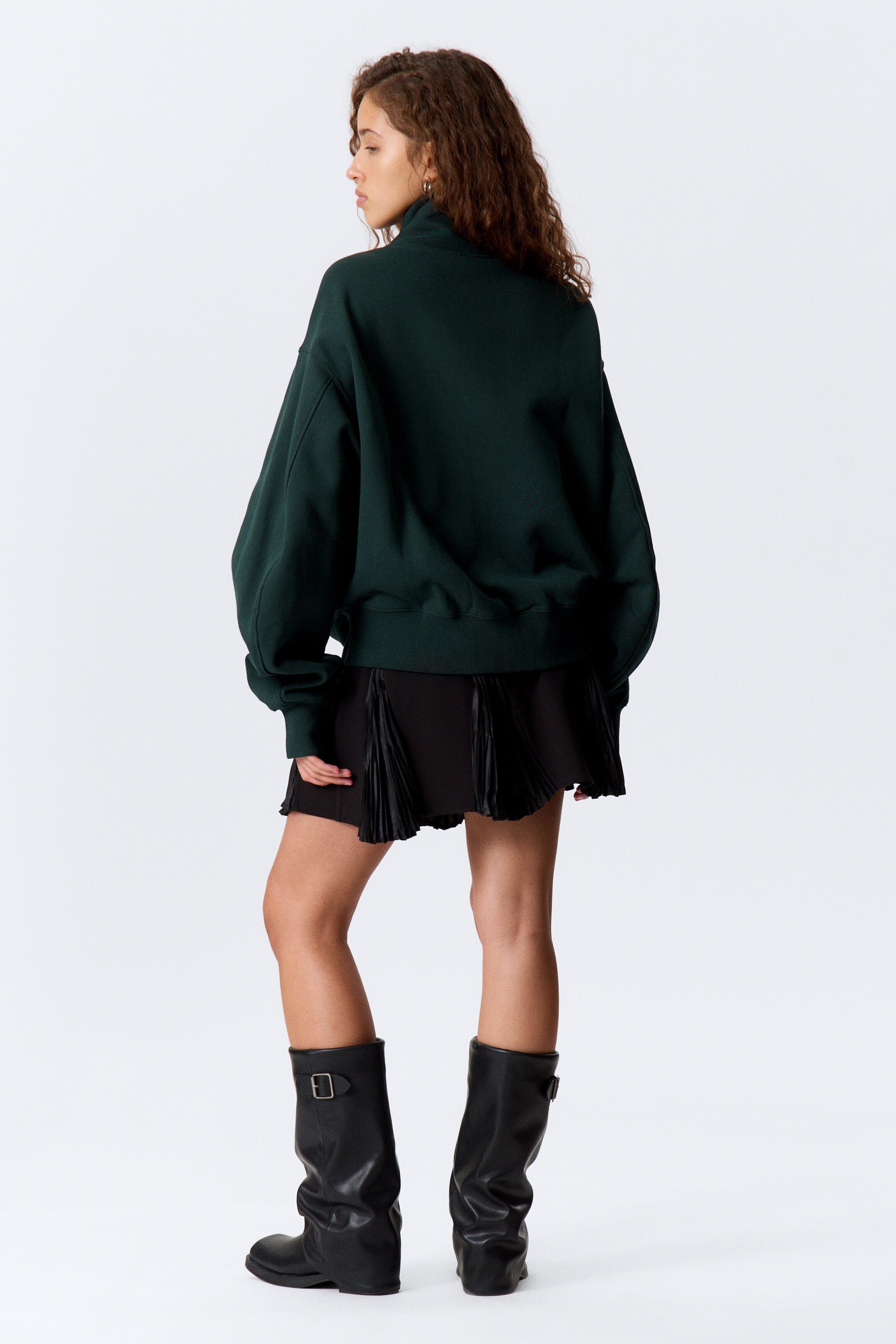 Visa större bild: En mörkgrön sweatshirt presenteras från baksidan på höger sida, och visar upp sin avslappnade passform. Den har en hög ståkrage, långa ärmar med ribbade muddar och en ribbad nederkant som faller precis ovanför en svart plisserad kjol.