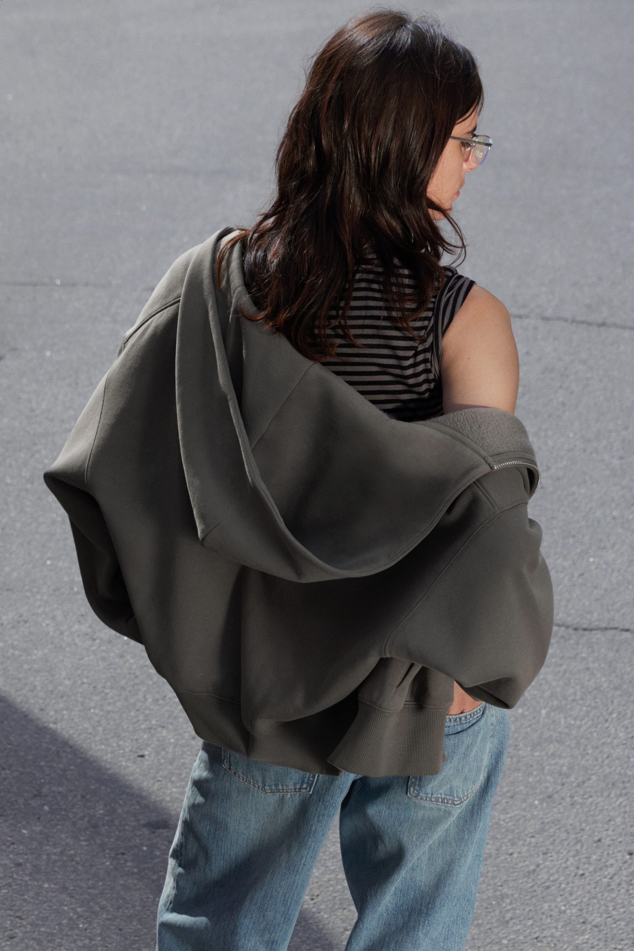 Agrandir l'image: Un sweat à capuche zippé intégral surdimensionné de couleur gris foncé est drapé sur les épaules, vu de dos. Il révèle un débardeur rayé noir et gris en dessous, et son ourlet côtelé repose au-dessus d'un jean bleu clair.