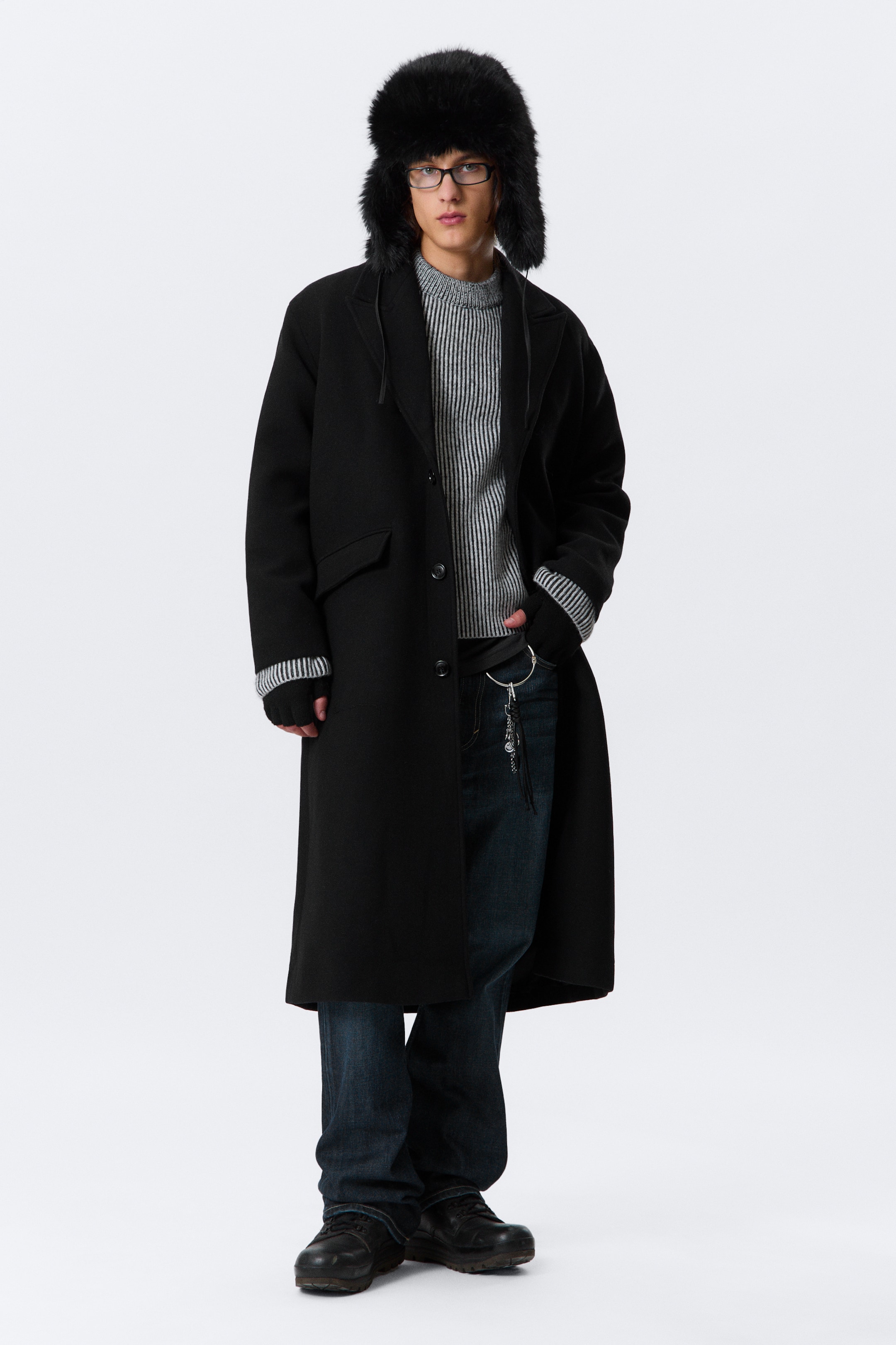 View larger image: A black long coat is being presented on a person facing forward. The oversized coat is featuring notched lapels, a three-button front closure, and two flap pockets. Its long sleeves are covering the arms, with striped sweater cuffs visibly extending from the wrists.