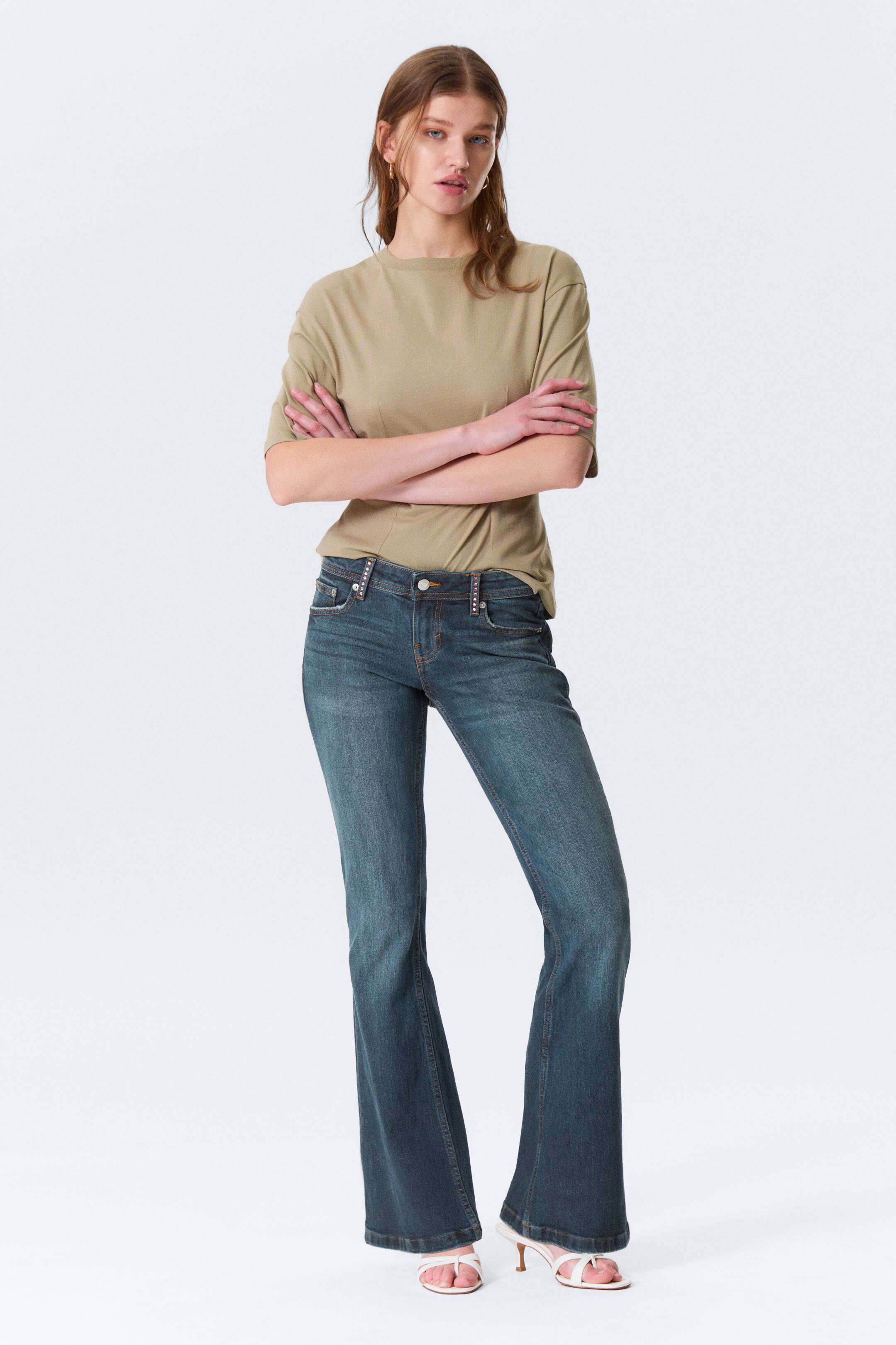 Zobrazit větší obrázek: Představujeme pár džínsů s vybledlým tmavě modrým praním, zobrazených zepředu, s přiléhavým střihem přes stehna a širokým rozšířením od kolen dolů. Džíny se vyznačují zapínáním na knoflík a zip, viditelnými předními kapsami a ozdobnými cvočky podél pasu.