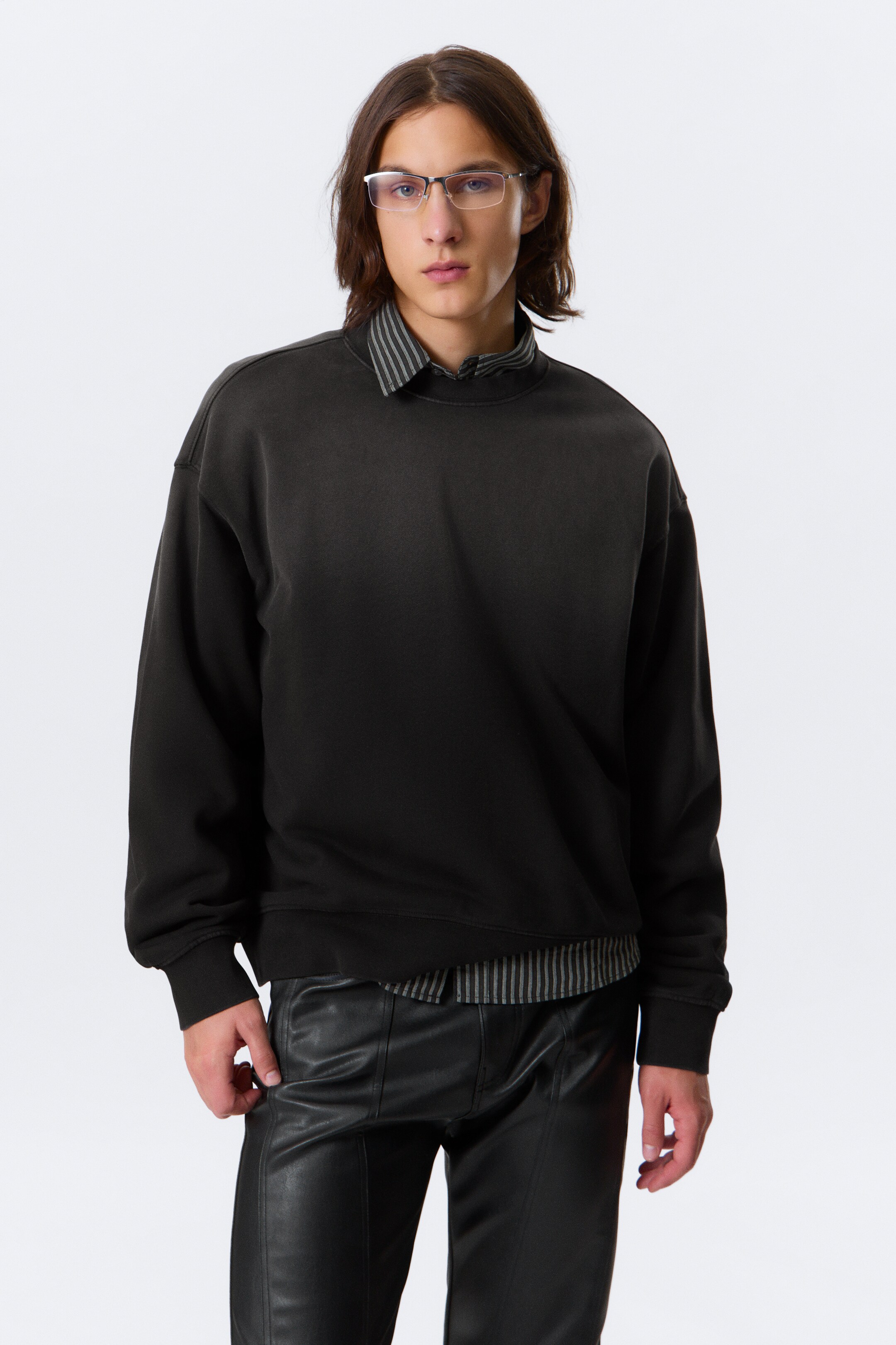 Grotere afbeelding bekijken: Een zwarte crewneck sweater wordt frontaal gepresenteerd, met een ontspannen pasvorm, lange mouwen, geribde boorden en een geribde zoom. De stof vertoont een gewassen, vervaagd effect, dat lichter lijkt over de borst en schouders, en een gestreept overhemd met kraag wordt eronder gelaagd met de kraag zichtbaar.