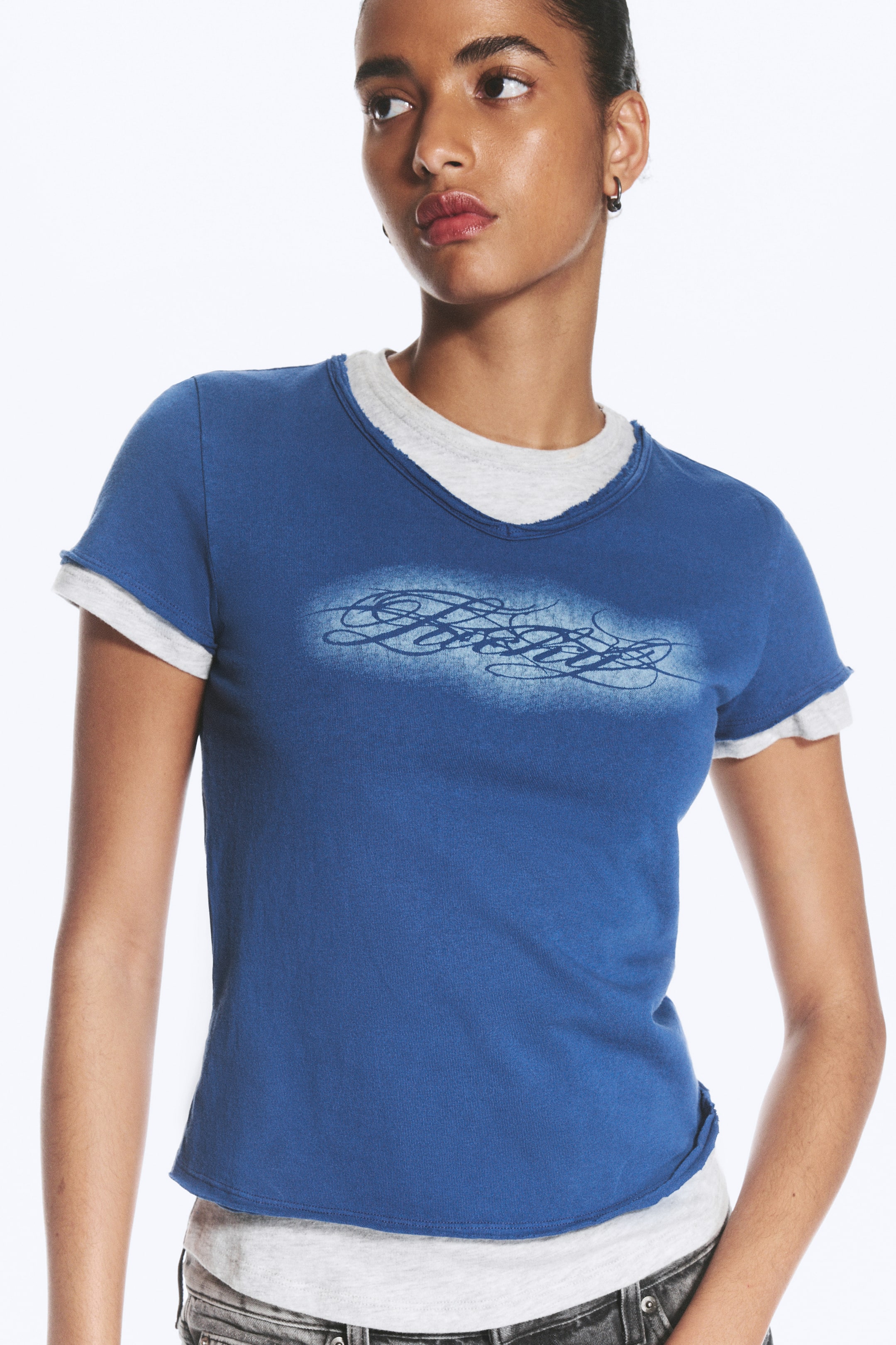 Visualizza immagine più grande: La modella indossa una T-shirt a doppio strato, caratterizzata da uno strato esterno a maniche corte blu con una stampa grafica sbiadita sul petto. Questo strato esterno è abbinato a una T-shirt interna grigio chiaro, visibile al girocollo con bordo grezzo, ai polsini delle maniche e all’orlo.