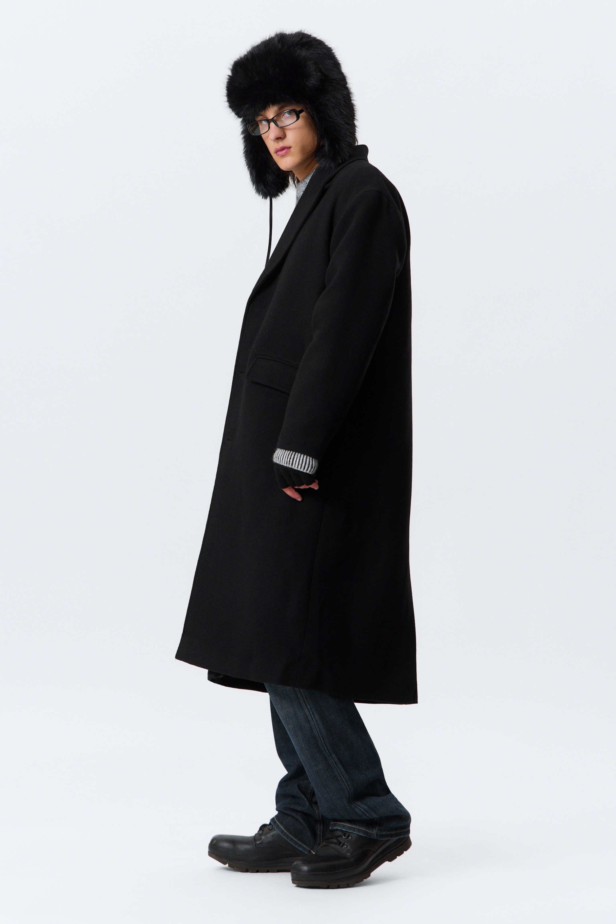 View larger image: A long black coat is being shown in a side profile, extending to just below the knees. It is featuring notch lapels and a flap pocket on the side. A gray and white striped cuff is peeking out from under the coat's sleeve.