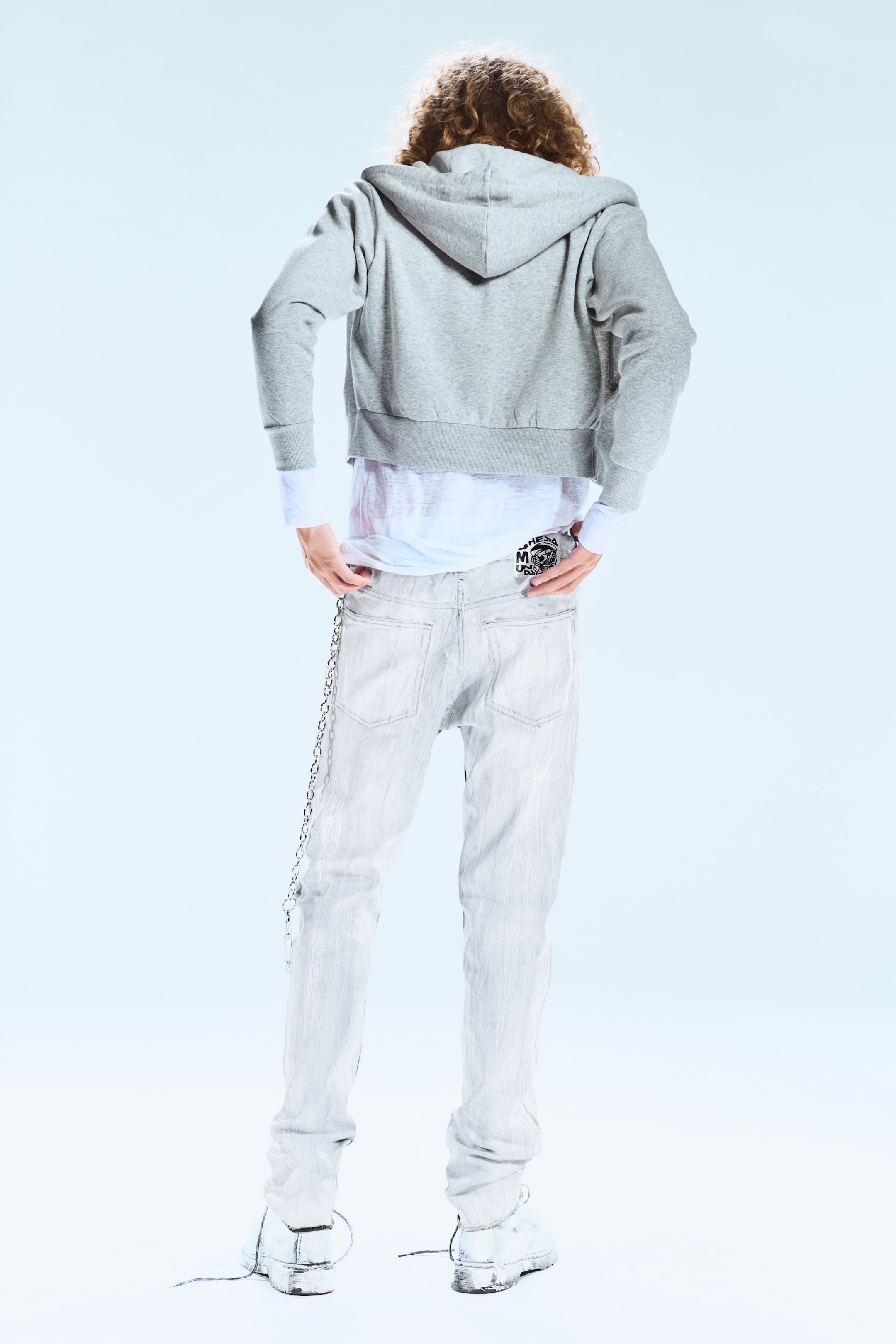 Agrandir l'image: Un sweat à capuche gris coupé est porté par-dessus une chemise blanche à manches longues. Un jean délavé clair, avec un détail de chaîne en métal sur le côté gauche et un patch graphique noir à l'arrière droit, est également porté par le mannequin, dont les mains reposent en haut des poches avant.